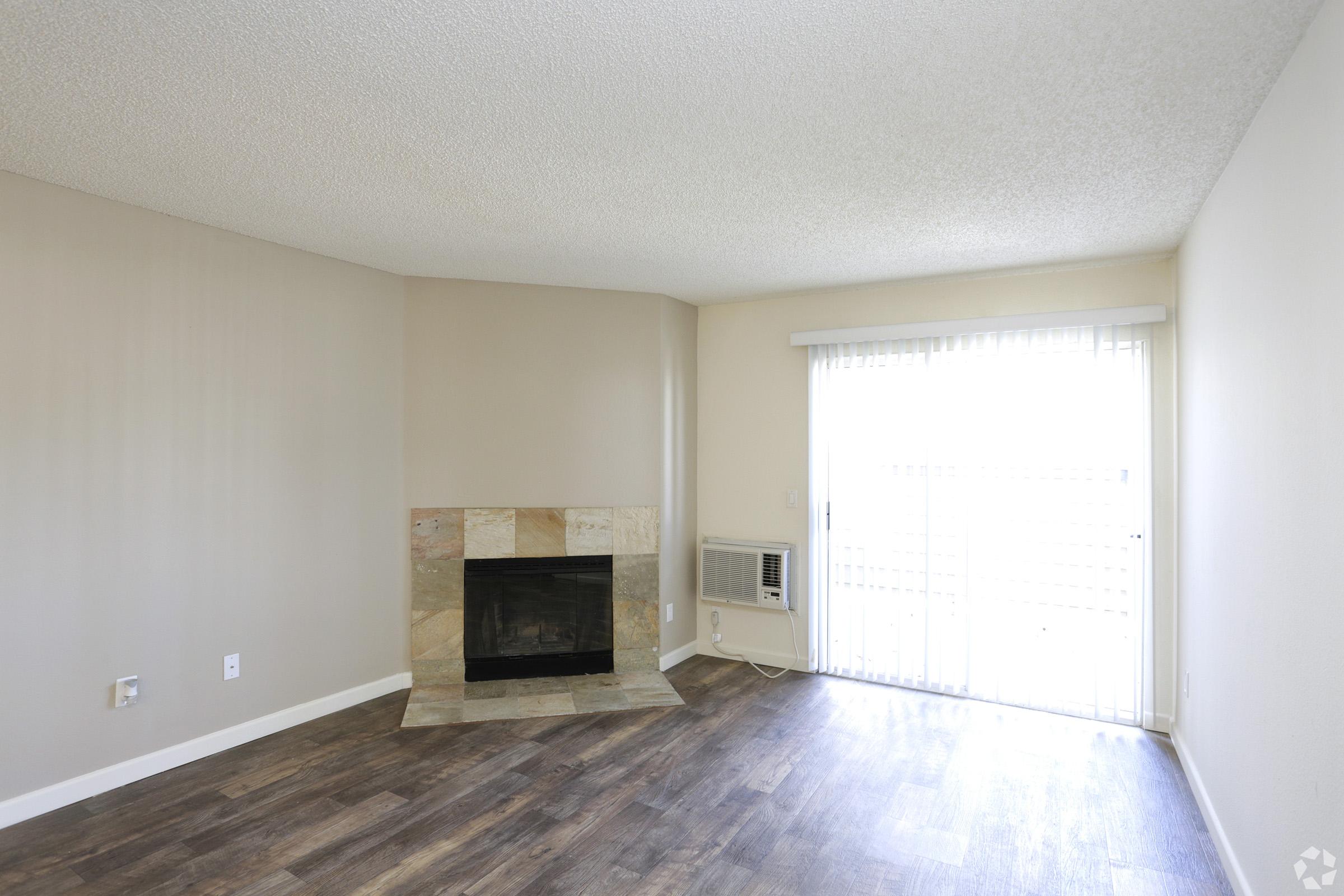 Interior of a living room featuring a stone fireplace, a sliding glass door with vertical blinds, and a wall-mounted air conditioning unit. The flooring is dark wood, and the walls are painted in light neutral colors, creating a spacious and inviting atmosphere.