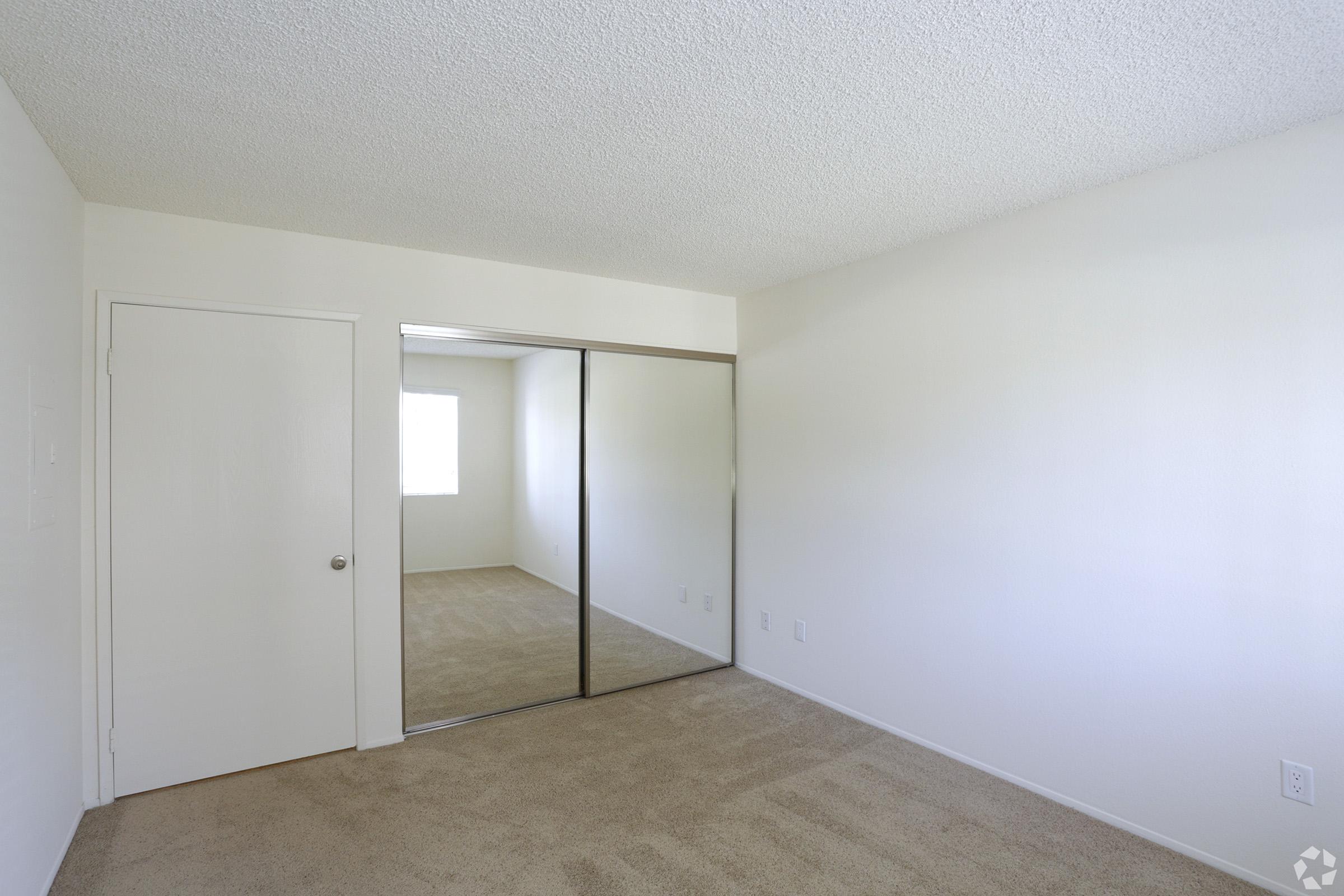 Empty room featuring light-colored walls and a soft carpet. There is a closed door on the left and a large mirrored closet on the right. A window allows natural light to enter, illuminating the space. The overall atmosphere is bright and minimalistic, suitable for various uses.