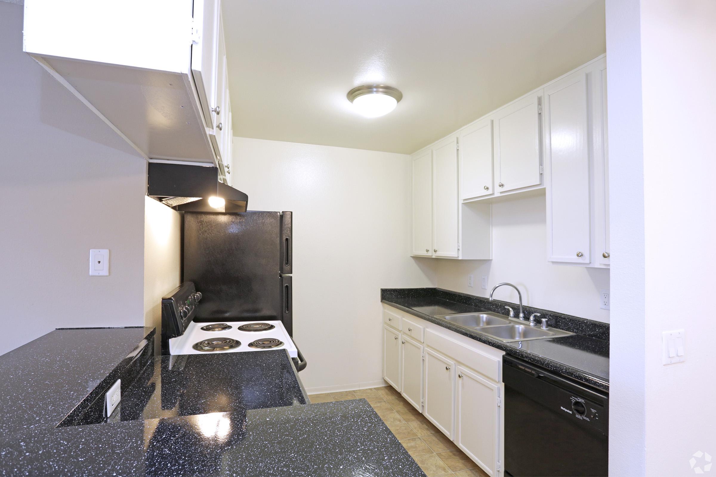 A modern kitchen featuring white cabinets, a black countertop, a stove with four burners, an oven, and a double sink. The walls are painted a light color, and there's a light fixture on the ceiling. The flooring is beige tile.