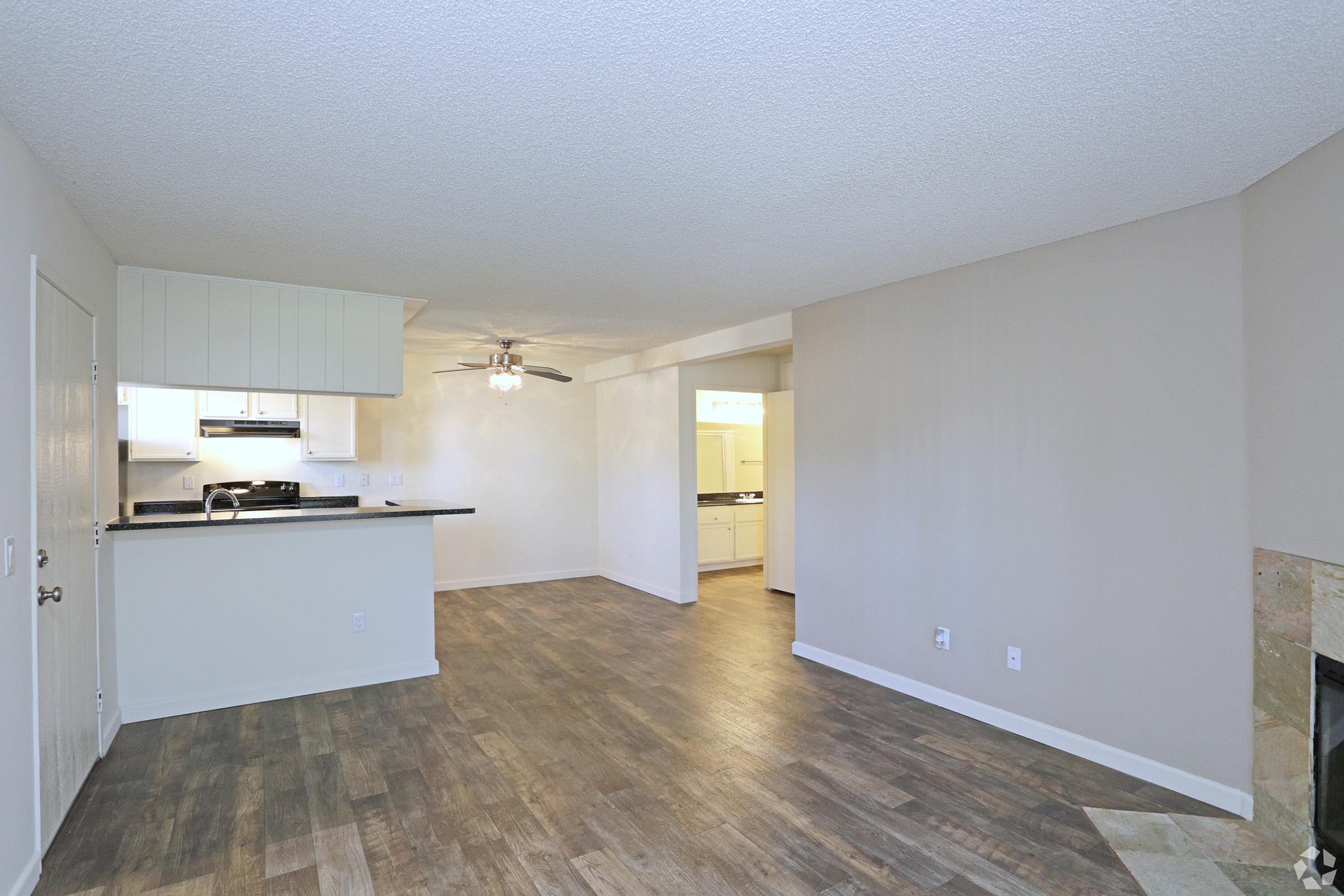 Spacious living area featuring wood-like flooring, a ceiling fan, and a fireplace. The open layout connects to a kitchen with modern appliances, visible in the background. Light-colored walls create a bright and inviting atmosphere, with a doorway leading to additional rooms.