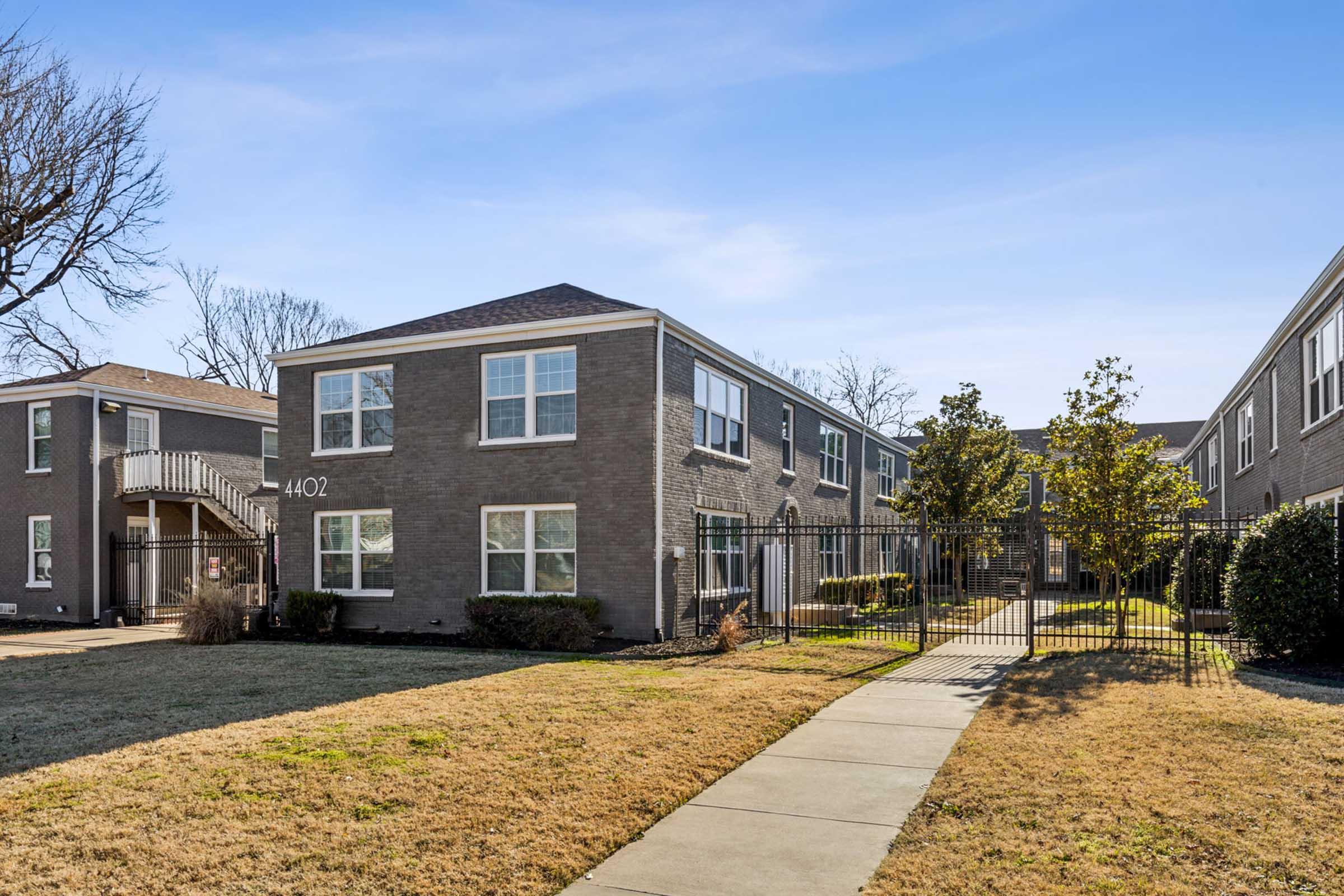 A well-maintained gray brick apartment building with multiple windows, featuring a fenced front yard and landscaped areas. A pathway leads to the entrance, and a fire escape is visible on the left side. Clear blue sky above, indicating a sunny day.