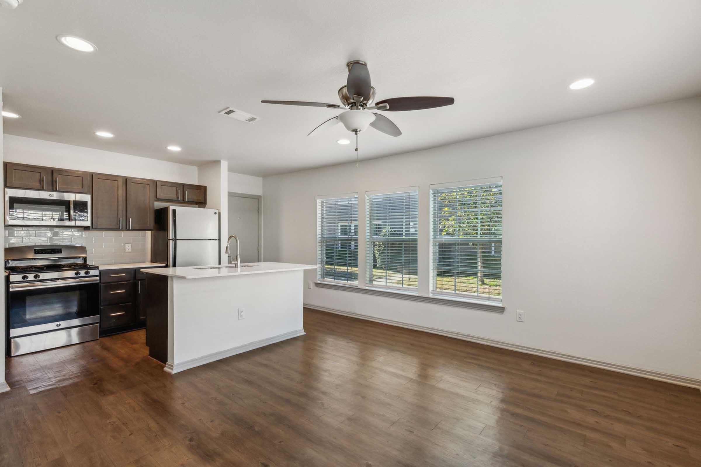 A modern kitchen and living area featuring stainless steel appliances, dark wooden cabinetry, and a central island. Large windows allow natural light to flood the space, highlighting the wood flooring. The room has a ceiling fan and a contemporary feel, with a view of the outdoor area.