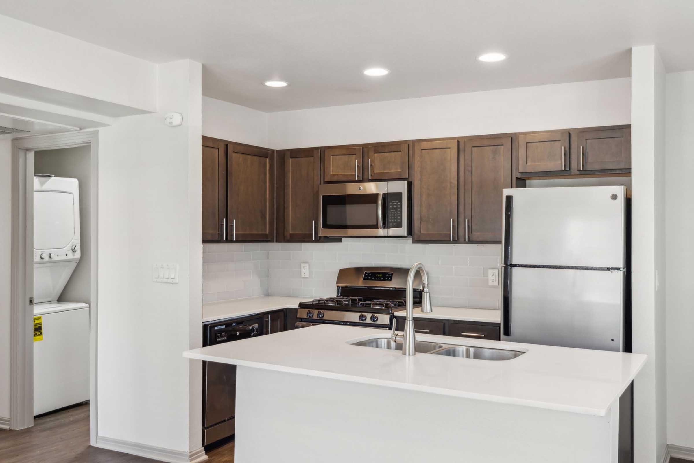 Modern kitchen featuring dark wood cabinets, stainless steel appliances, and a white countertop. The kitchen includes a gas stove, microwave, and refrigerator. Bright LED lighting illuminates the space, which has a sleek design and a functional layout. A laundry area is visible in the background.