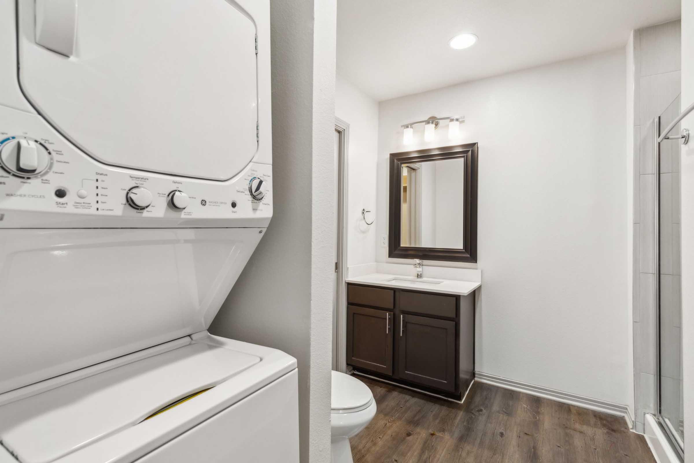 A modern bathroom featuring a stacked washer and dryer, a vanity with a mirror, a toilet, and a glass shower. The space has neutral-colored walls and a wooden floor, providing a clean and functional design.