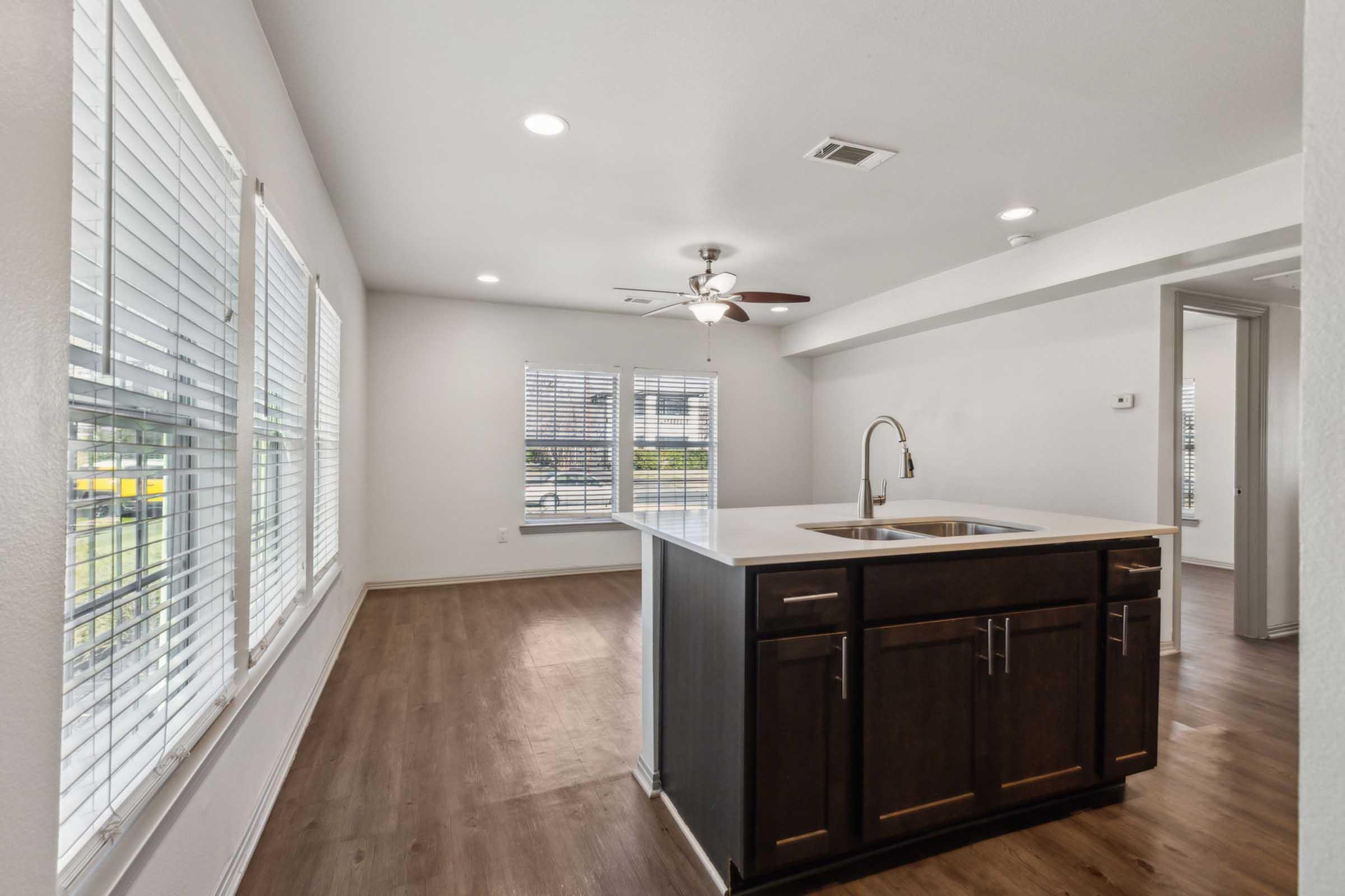 Modern, well-lit interior of a kitchen and living area featuring a central island with a sink, dark cabinetry, and large windows with blinds. The space has a clean, minimalist design with neutral wall colors and wooden flooring, creating an inviting atmosphere.