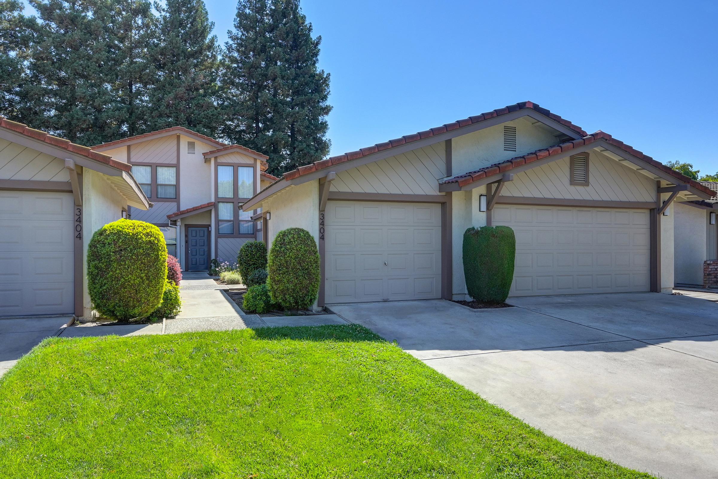 A residential area featuring two single-story townhouses with attached garages. Lush green lawns and neatly trimmed hedges are in front of the homes. In the background, a larger two-story home can be seen, surrounded by tall trees under a clear blue sky.