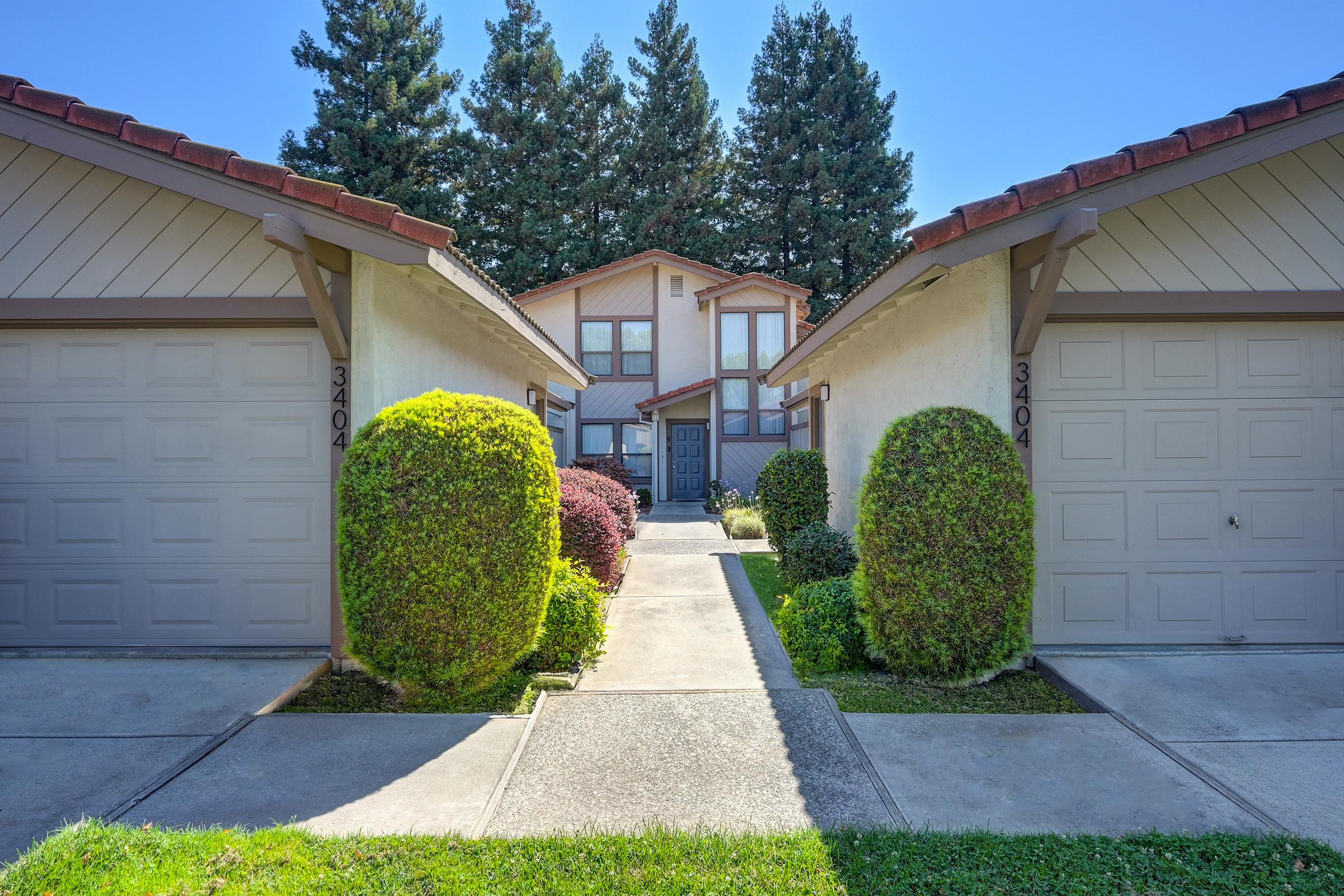 A landscaped pathway flanked by two identical residential buildings, each featuring a single garage door. Lush greenery and neatly trimmed hedges enhance the entrance, with a clear blue sky overhead. The buildings have a light exterior color and a brown tiled roof, creating a welcoming atmosphere.