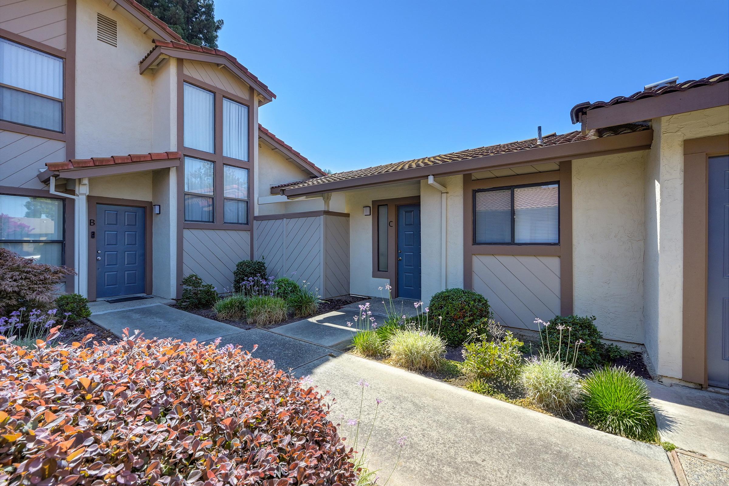 A view of a residential building entrance featuring a gray front door surrounded by neatly landscaped plants and flowers. The architecture includes light-colored walls and a tiled roof, with a clear blue sky in the background. The pathway is lined with greenery, enhancing the inviting atmosphere.