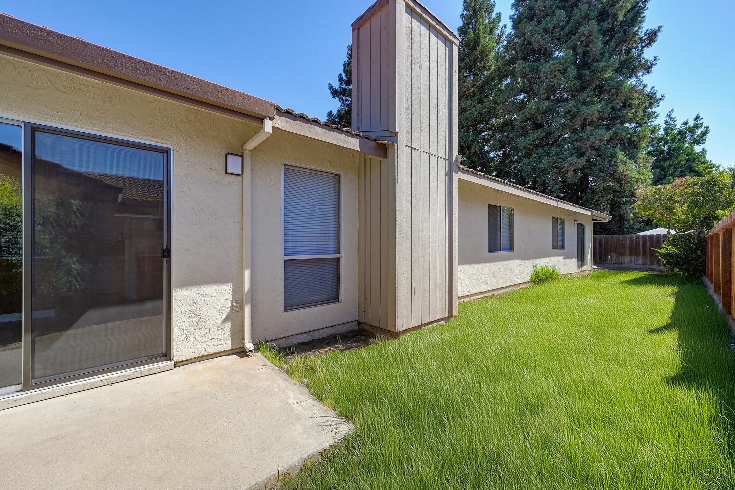 A side view of a residential house featuring a small lawn with green grass. The house has a light-colored exterior with large windows and a tall chimney. The background includes trees and a wooden fence, providing a sense of privacy. Clear blue skies indicate a sunny day.