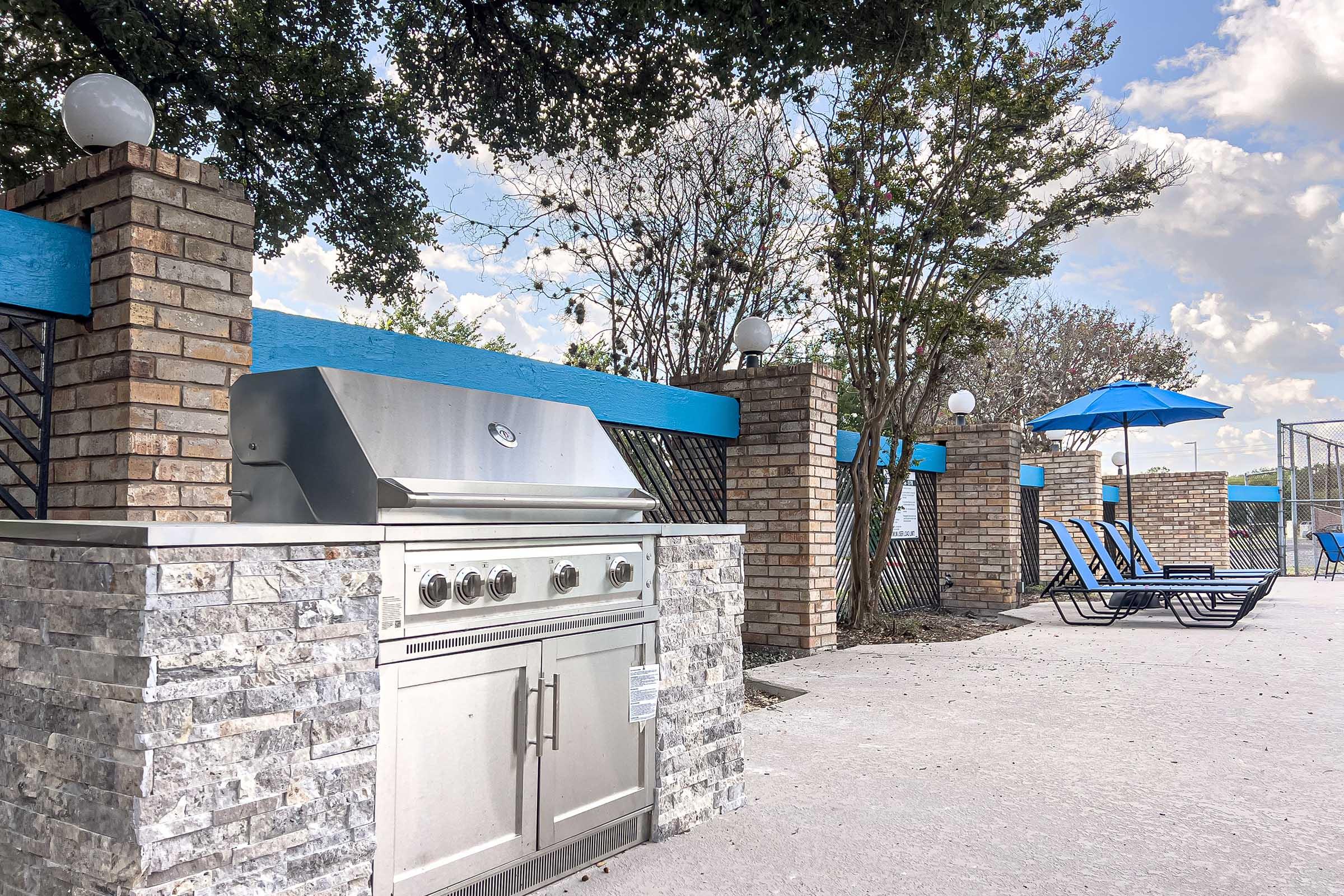A modern outdoor grill made of stone and stainless steel is situated beside a pool area. Blue lounge chairs are arranged nearby, with a large umbrella providing shade. Lush trees and a clear blue sky with fluffy clouds complete the scene, inviting relaxation and outdoor cooking.