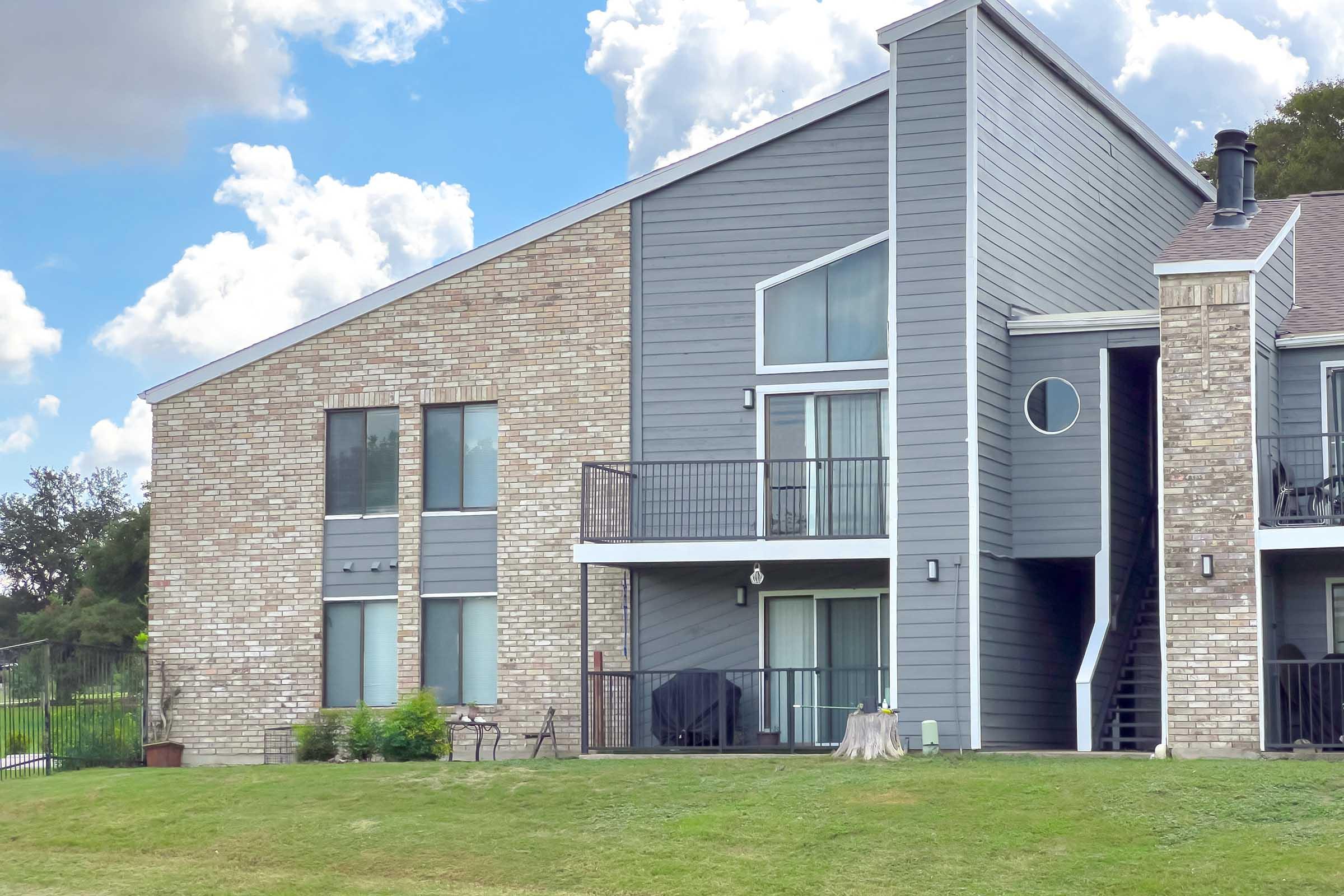 Two-story apartment building featuring a combination of brick and gray siding, large windows, and balconies. The exterior includes a small grassy area in the foreground, with a tree stump, and a fence visible in the background. The sky is partly cloudy.