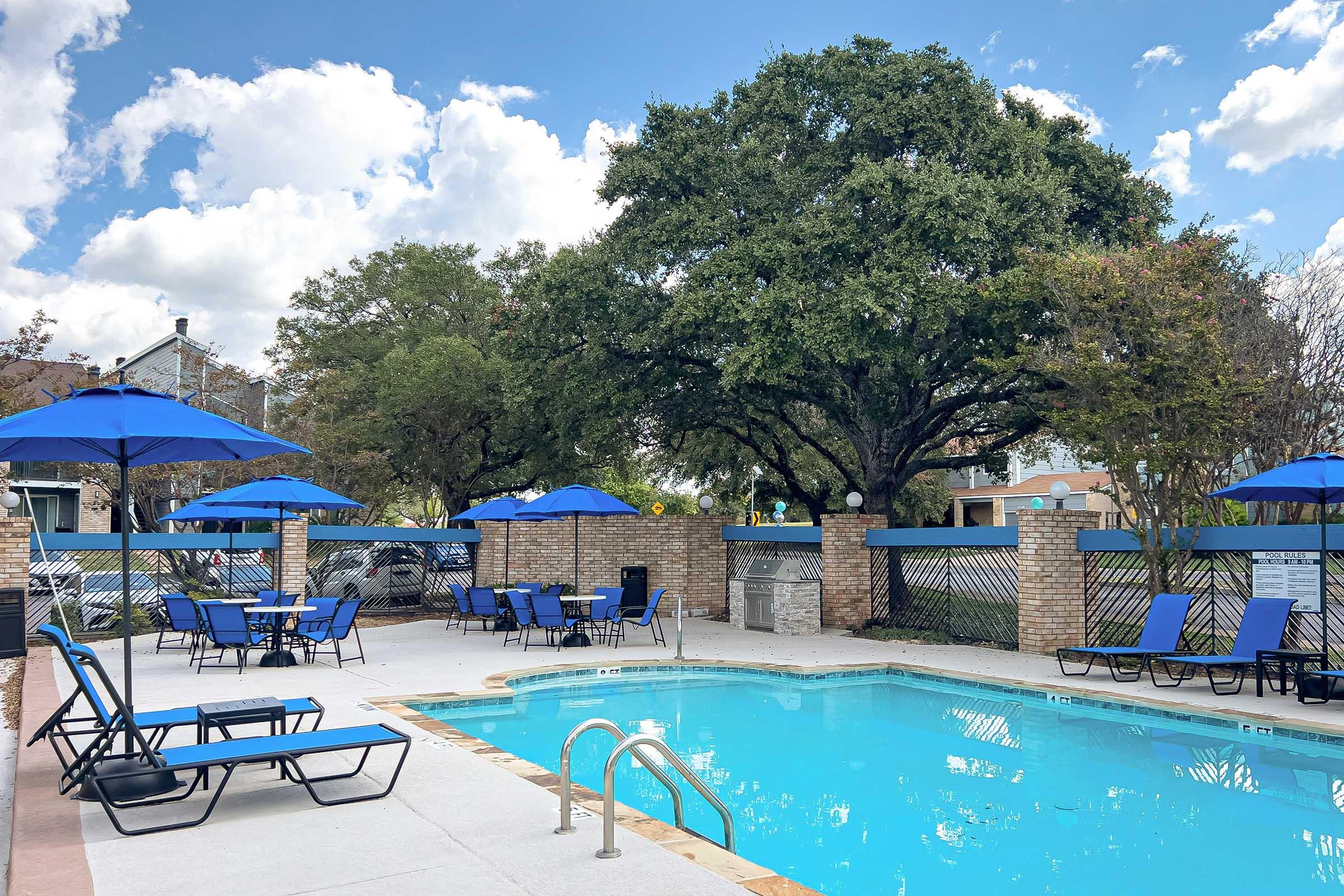 A serene swimming pool area featuring a clear blue pool surrounded by lounge chairs and blue umbrellas. Lush trees and a brick wall create a tranquil atmosphere, with picnic tables and a grill nearby. Bright clouds float in the blue sky, enhancing the inviting outdoor space.