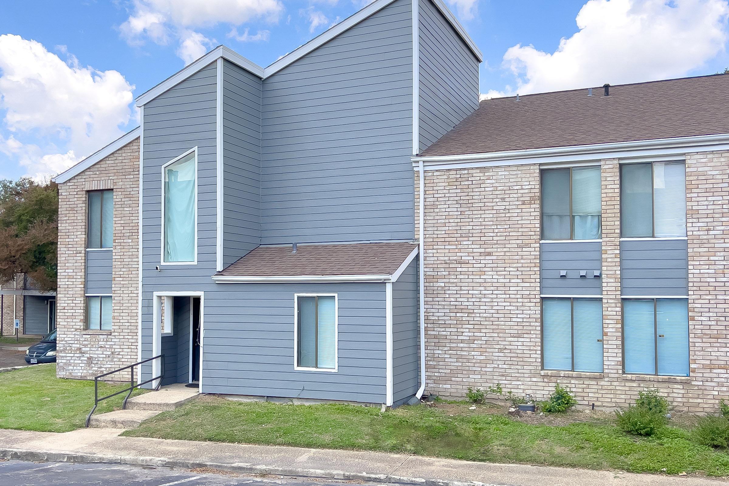 A modern multi-unit residential building featuring a combination of brick and gray siding. The structure has a distinctive angular design with large windows. A small front porch with a railing leads to the main entrance, and the surrounding area includes a grassy lawn and a driveway.
