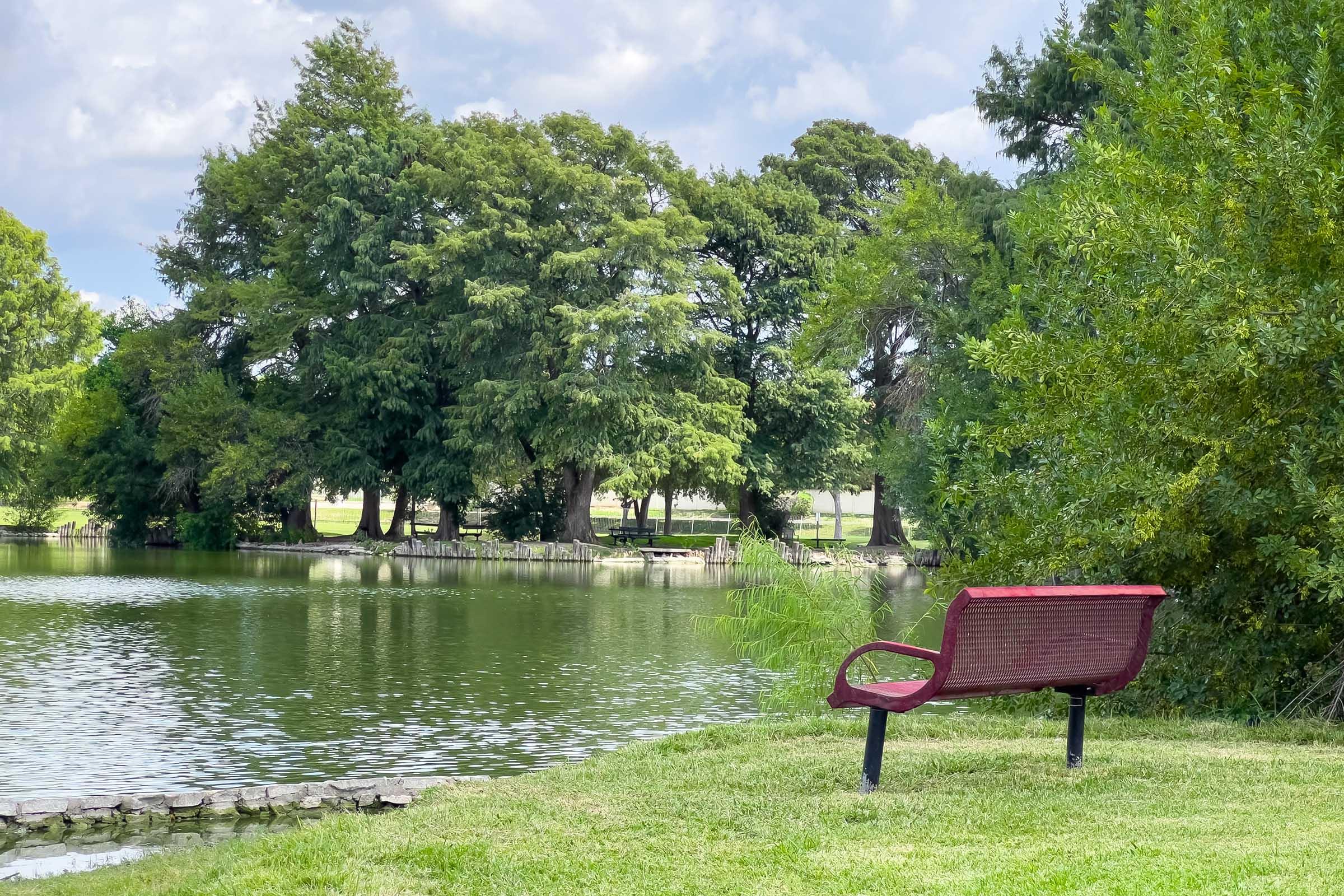 A serene pond surrounded by lush greenery and trees, with a red bench positioned near the water's edge. The sky is partly cloudy, creating a peaceful outdoor scene ideal for relaxation and enjoying nature.