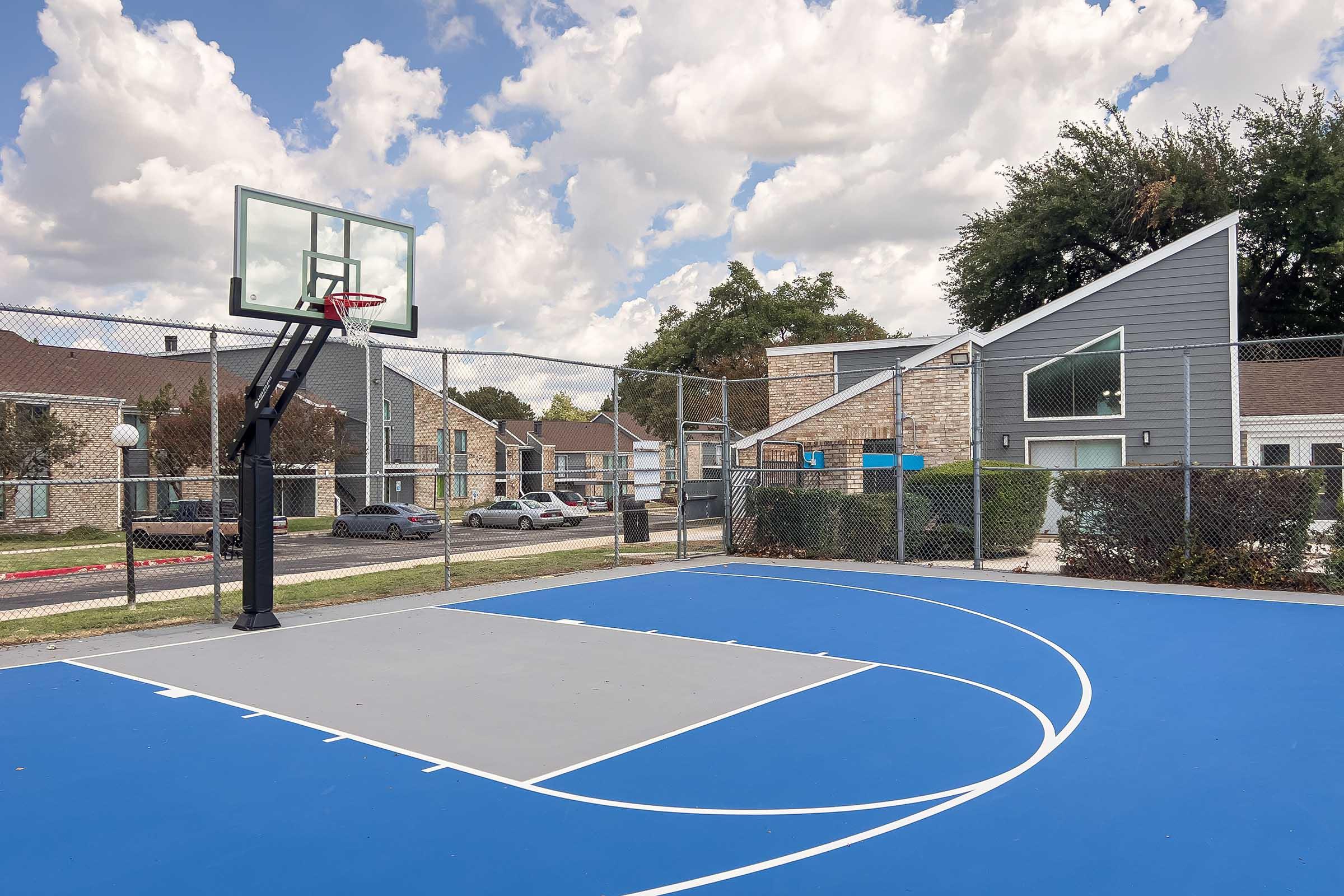 A basketball court with a blue surface, surrounded by a chain-link fence. In the background, there are residential buildings and a cloudy sky. The court features a backboard and hoop, with parking visible near the buildings.