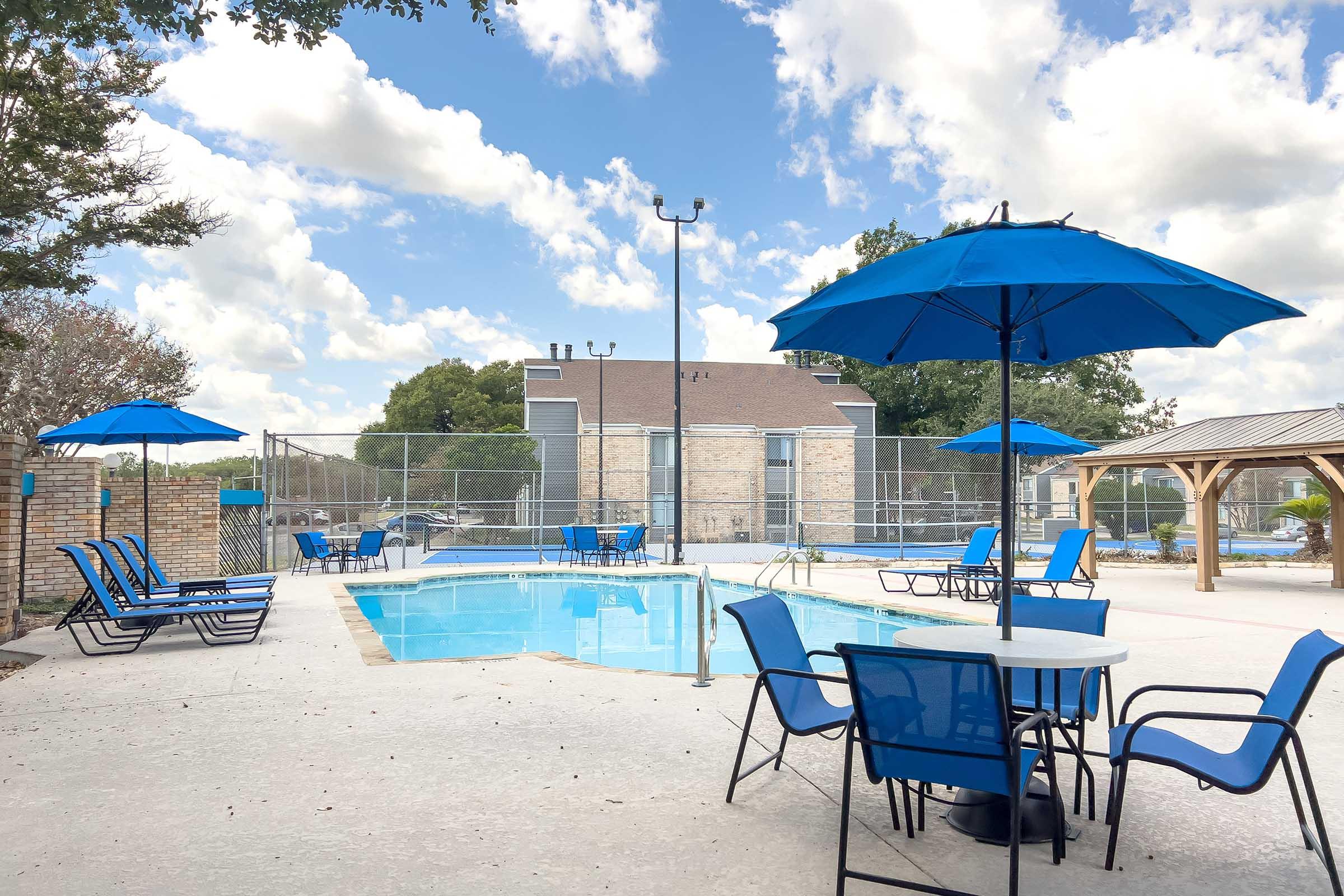 A serene outdoor pool area featuring a clear blue pool surrounded by lounge chairs and tables with vibrant blue umbrellas. Lush trees provide a natural backdrop, and a fenced area adds privacy. The sky is partly cloudy, creating a pleasant atmosphere for relaxation.