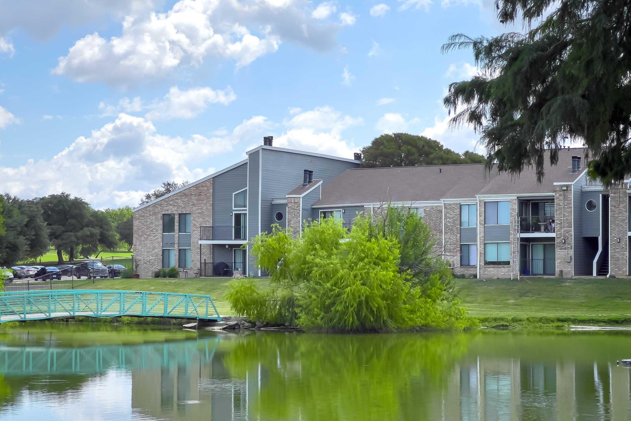 A serene view of an apartment complex beside a calm pond, featuring lush green trees and a small turquoise bridge crossing over the water. The building has a mix of stone and modern architecture, with large windows reflecting the surrounding greenery under a partly cloudy sky.