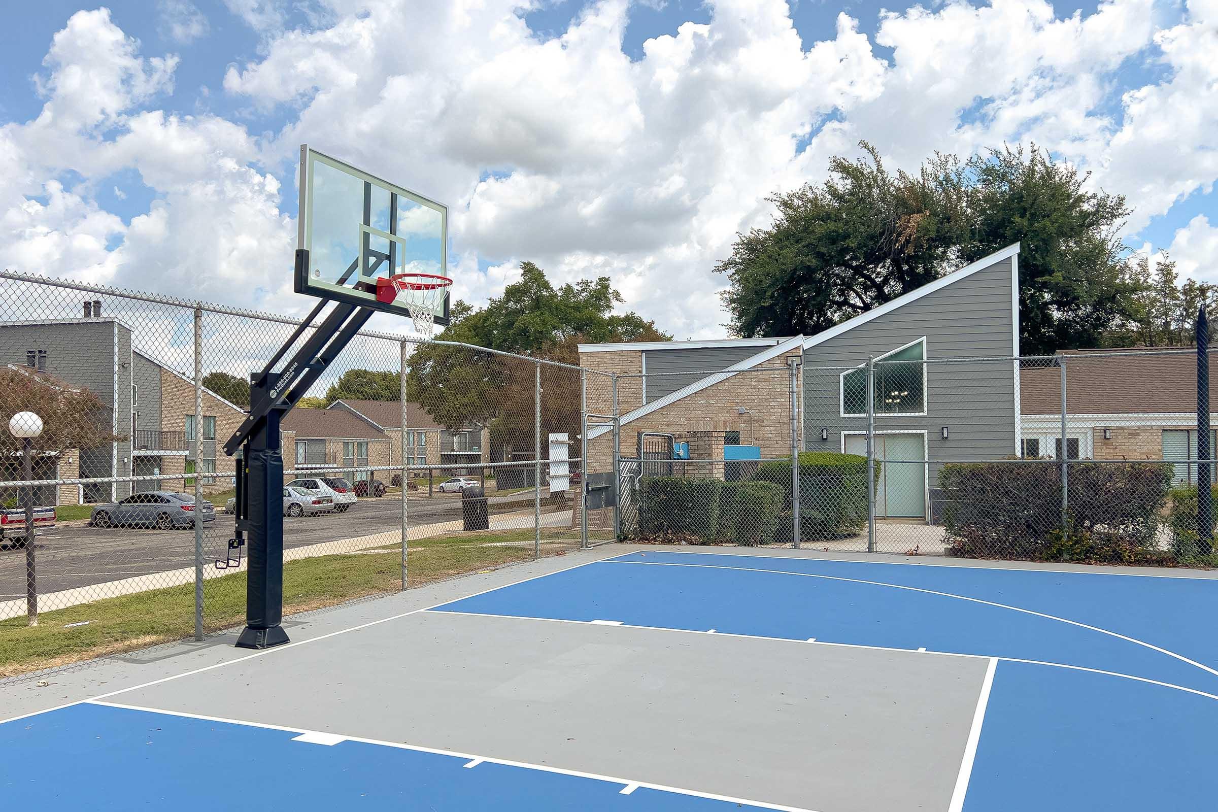 A basketball court with a hoop on a sunny day. The court features blue and grey surfaces, surrounded by a chain-link fence. In the background, there are residential buildings and a tree-lined area under a partly cloudy sky.