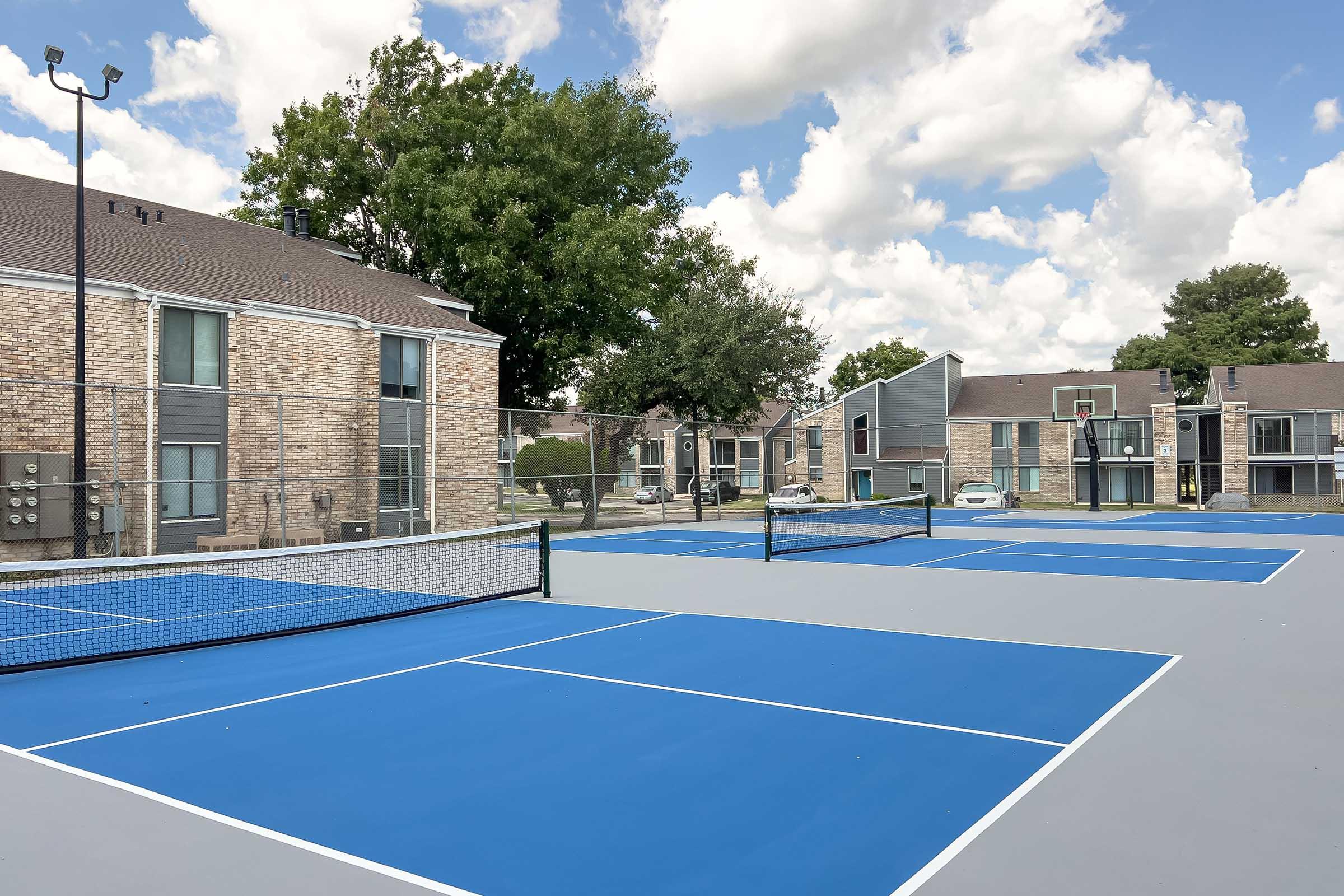Empty outdoor tennis courts with blue surfaces and black nets, surrounded by residential buildings. The sky is partly cloudy with a few trees in the background, creating a bright and inviting atmosphere.