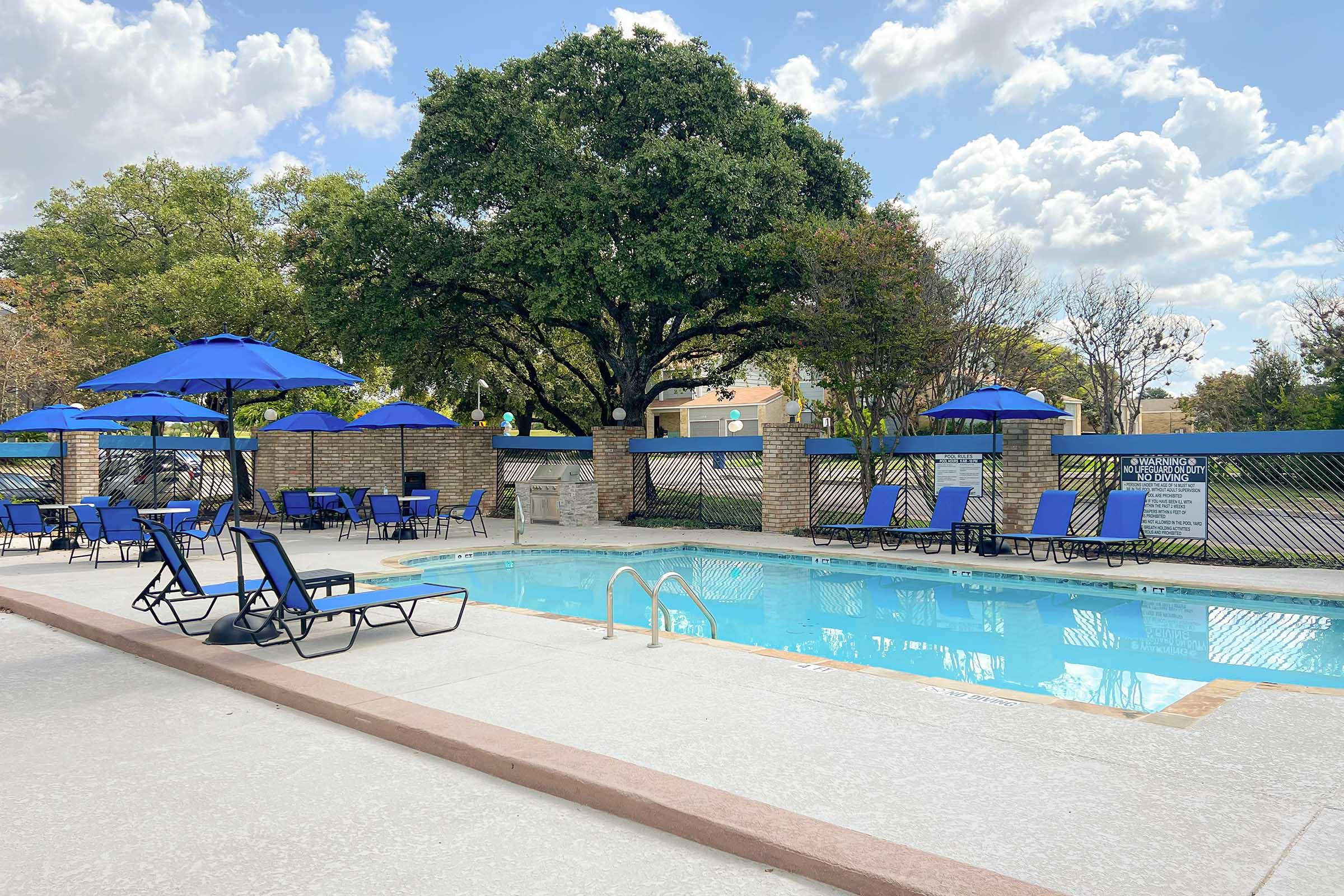 A serene pool area featuring a clear blue swimming pool surrounded by lounge chairs with blue umbrellas. Lush trees and a cloudy sky add to the relaxing atmosphere, making it an inviting space for leisure and recreation.