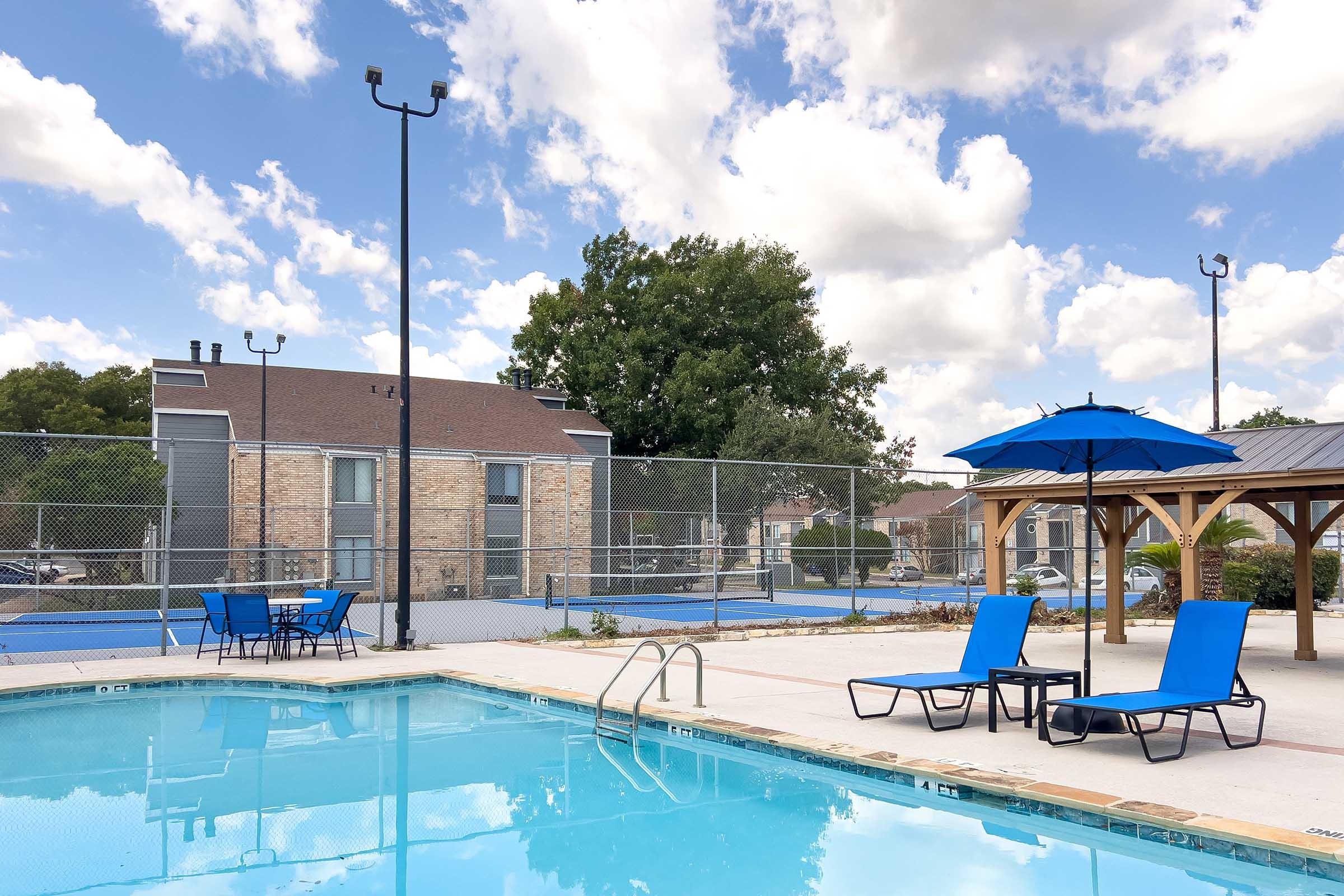 A refreshing swimming pool with blue lounge chairs and an umbrella, surrounded by a fenced area. In the background, two tennis courts and an apartment complex are visible under a partly cloudy sky.