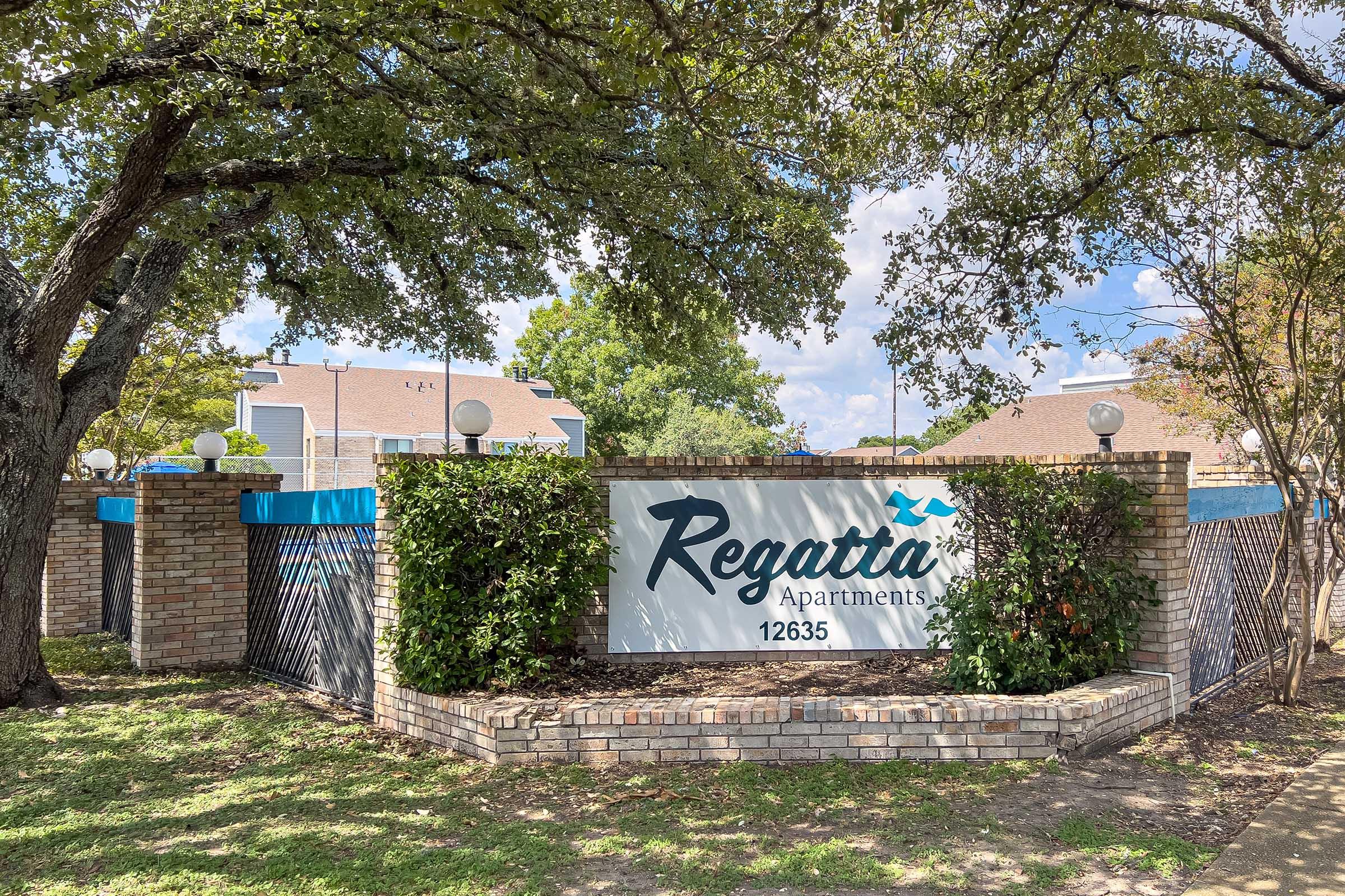 A stone sign featuring the name "Regatta Apartments" surrounded by greenery and trees, with a blue trim at the top. The sign is located in front of an apartment complex, with visible buildings and a clear sky in the background.