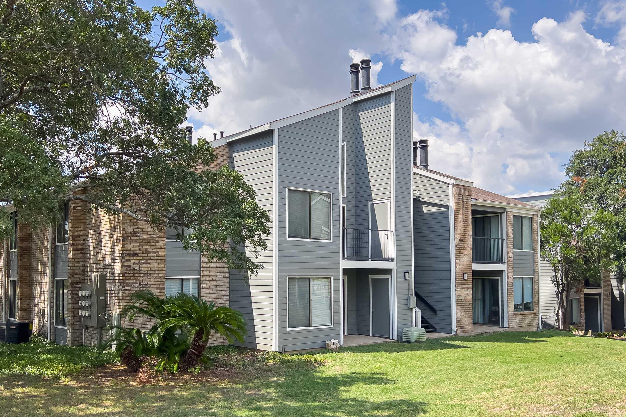 A multi-story apartment building featuring a mix of brick and gray siding. The building has large windows, a balcony, and is surrounded by green grass and trees. Partly cloudy sky in the background.