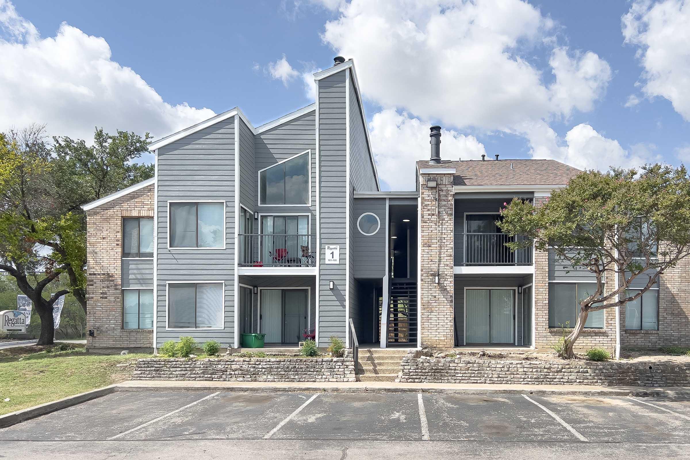 A modern two-story apartment building featuring a mix of brick and gray siding. The building has large windows, a circular architectural feature, and a small patio area. Surrounding greenery and a parking lot are visible in the foreground under a partly cloudy sky.