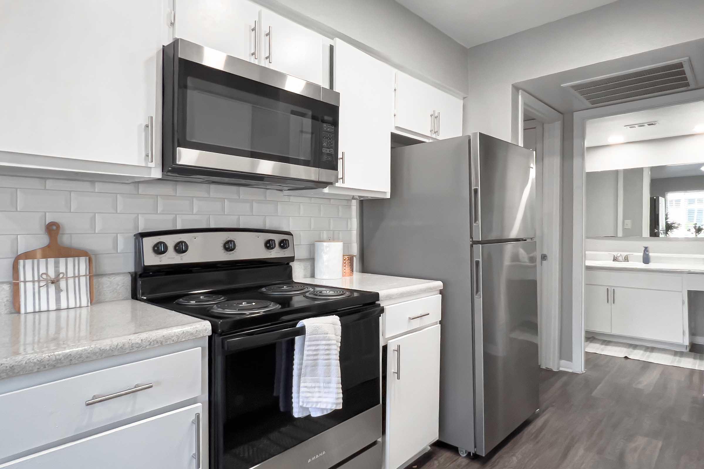 A modern kitchen featuring a stainless steel microwave above a black electric stove and oven. The cabinets are white with sleek handles, and there's a silver refrigerator visible. The countertop is light gray, and a white dish towel hangs from the oven. A mirror and sink area can be seen in the background.