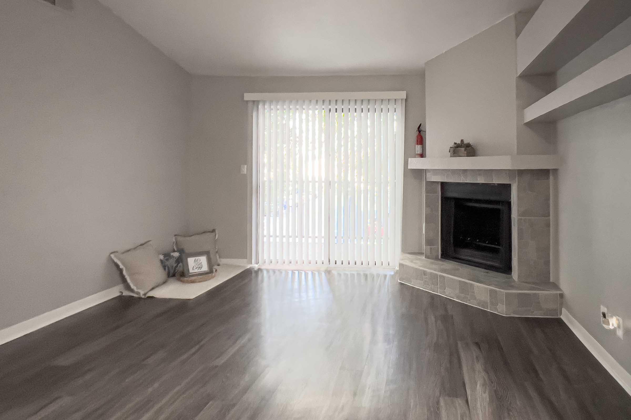 A spacious, modern living room with light gray walls and dark wood flooring. Large vertical blinds cover a glass door that lets in natural light. To the left, there are two soft cushions on the floor, and on the right, a stone fireplace sits in the corner, adding a cozy touch to the decor.
