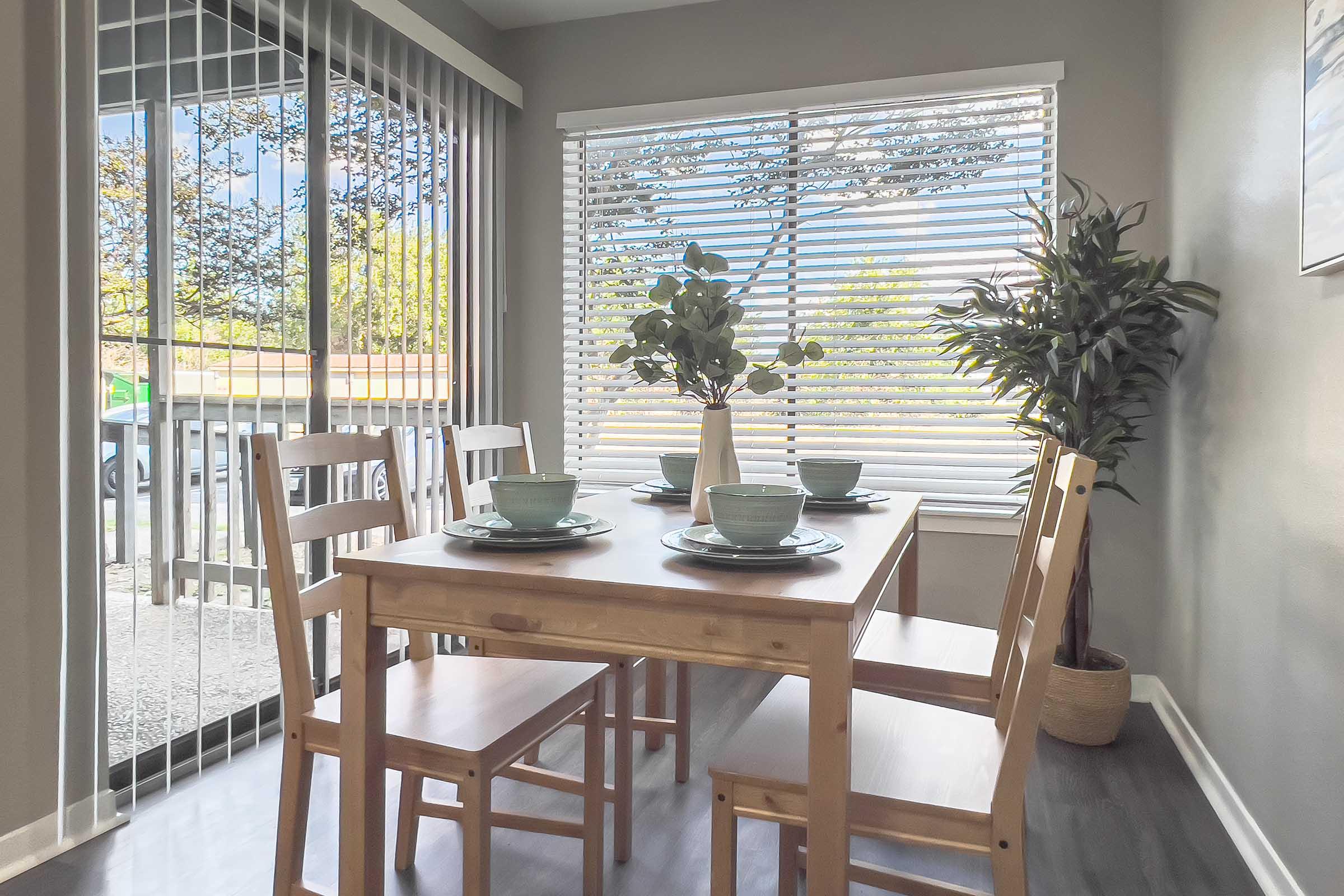 A cozy dining area featuring a wooden table set for four with light blue dishes and cups. A vase with greenery sits in the center. Sunlight streams through large windows adorned with white blinds, and there's a potted plant in the corner, creating a warm and inviting atmosphere.