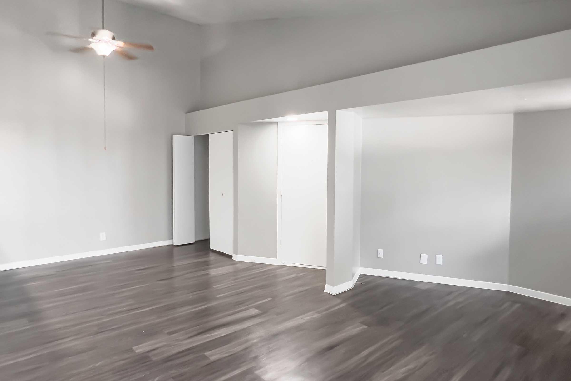 A spacious indoor area featuring light gray walls and dark wood-like flooring. A ceiling fan is mounted in the upper left corner. There are two closets with white doors on the left and an open wall archway leading to another room on the right. Natural light illuminates the space, creating a bright and airy atmosphere.