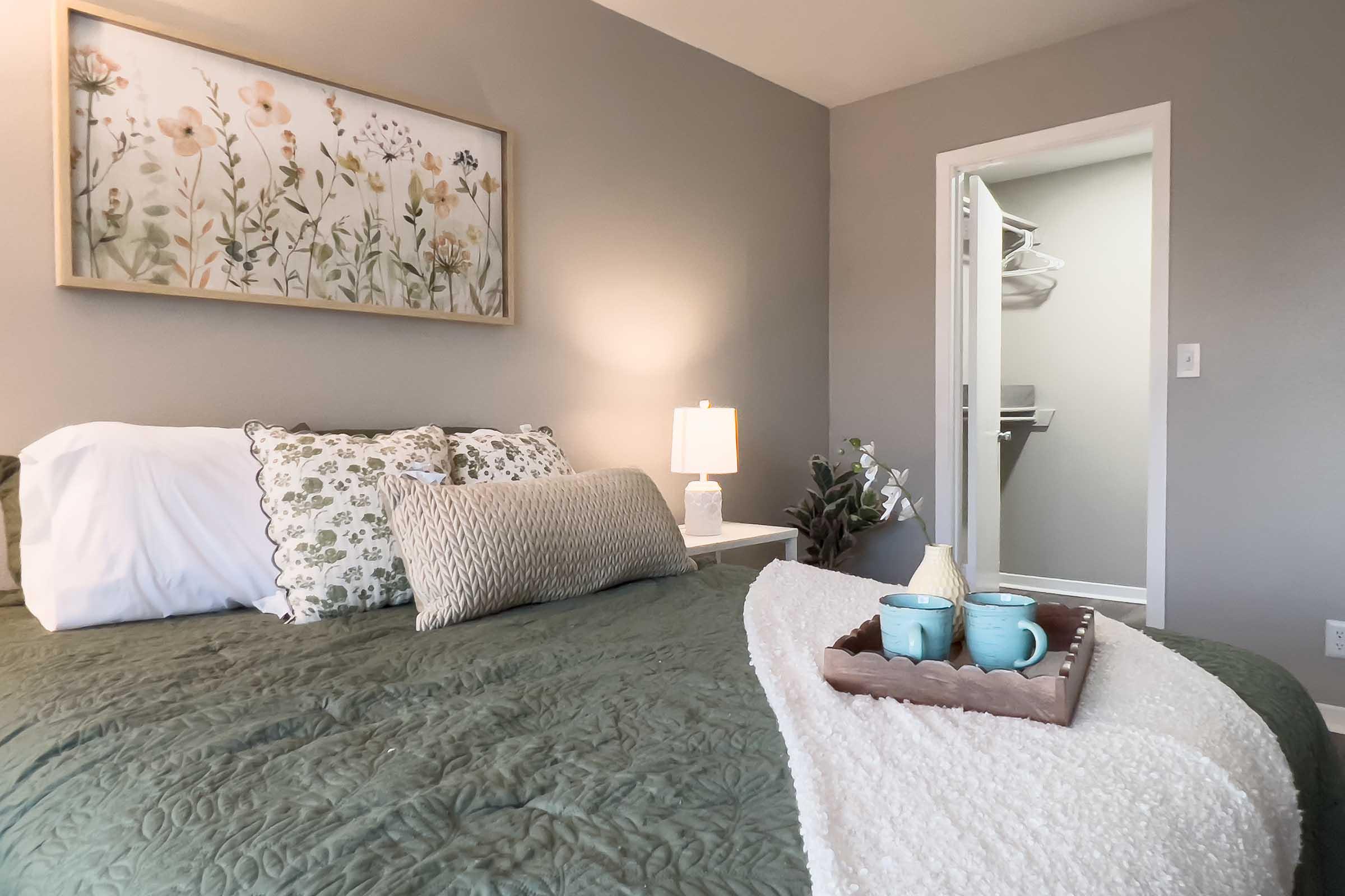 A cozy bedroom featuring a green quilted bedspread adorned with decorative pillows. A nightstand with a lamp and a tray holding two blue mugs sits beside the bed. A floral artwork hangs on the wall, and a nearby open closet reveals additional space. The walls are painted in a calming gray tone, enhancing the serene atmosphere.