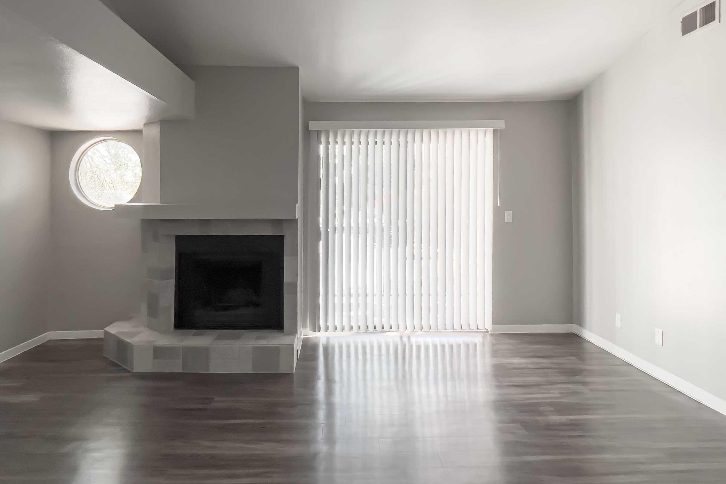 A spacious, modern living room featuring a circular window and a stone fireplace. The walls are painted light gray, and there are sliding glass doors covered with vertical blinds, allowing natural light to brighten the room. The floor is polished hardwood, creating a contemporary and inviting atmosphere.