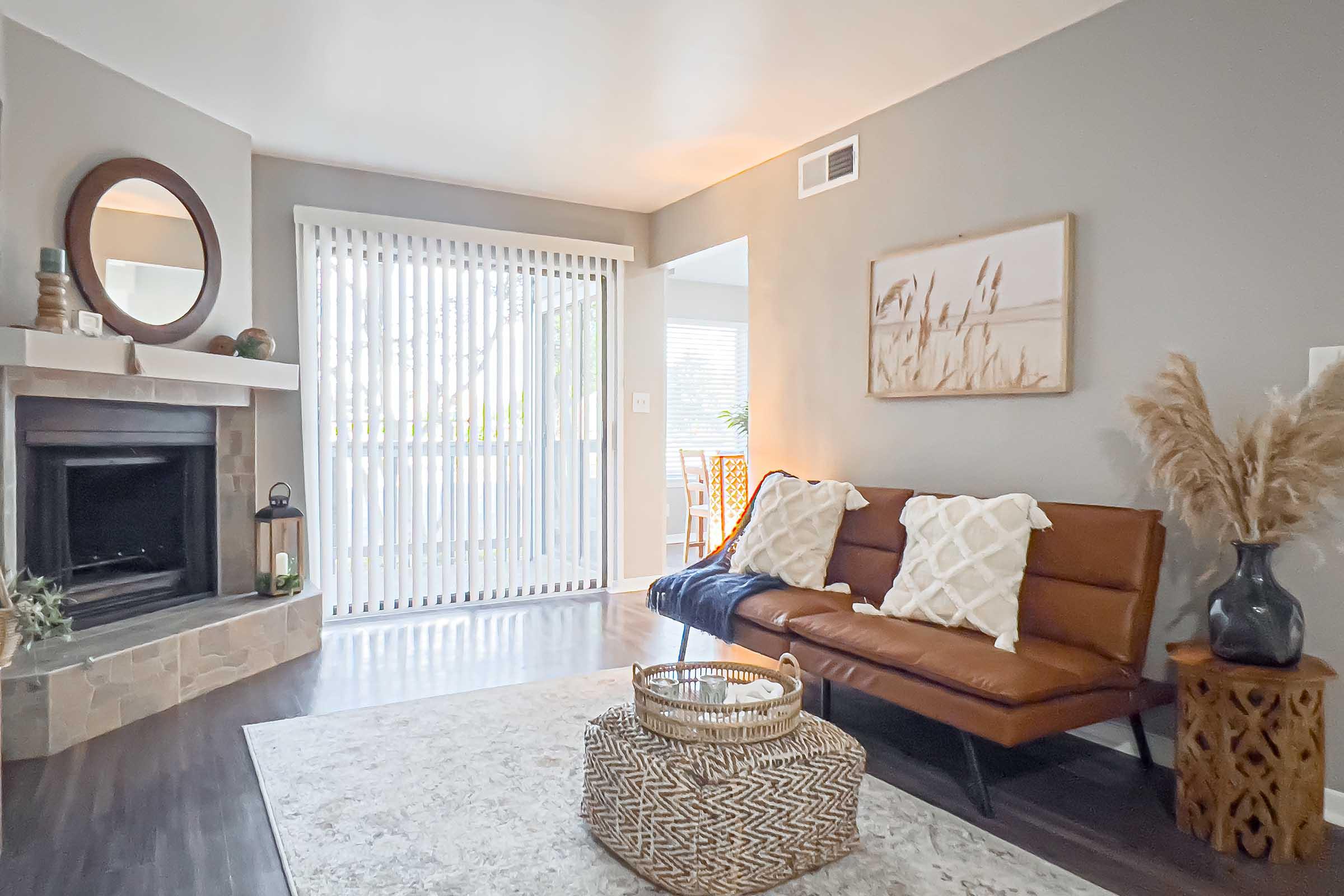 A cozy living room featuring a modern brown sofa with white cushions, a decorative area rug, and a stone fireplace. A round mirror hangs above the fireplace, and large sliding doors with blinds lead to an outdoor area. A basket and potted plants add natural elements to the serene atmosphere.