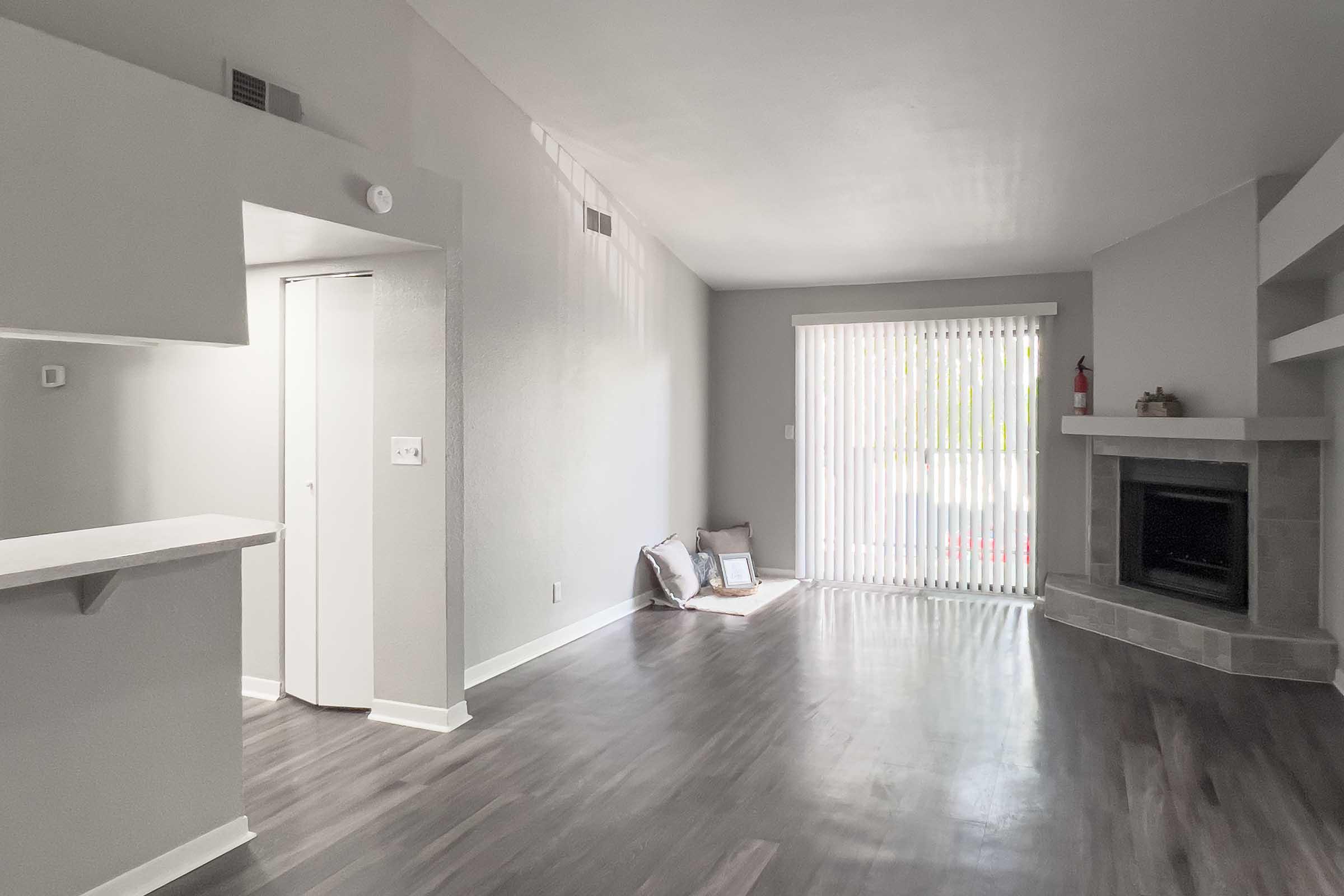 Spacious living area featuring gray walls and dark wood flooring. A large window with vertical blinds lets in natural light, and a fireplace is visible on the right. There's an open doorway leading to another room, and cushions are placed near the window for a cozy touch.