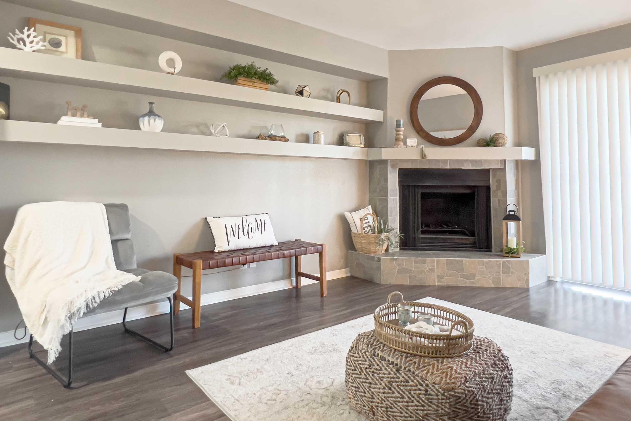 A cozy living room featuring a stone fireplace, gray accent walls, and a wood shelf with decorative items. There is a light gray chair, a wooden bench with a decorative pillow that says "Welcome," a round mirror above the fireplace, and a woven basket on the floor, complemented by a soft area rug.