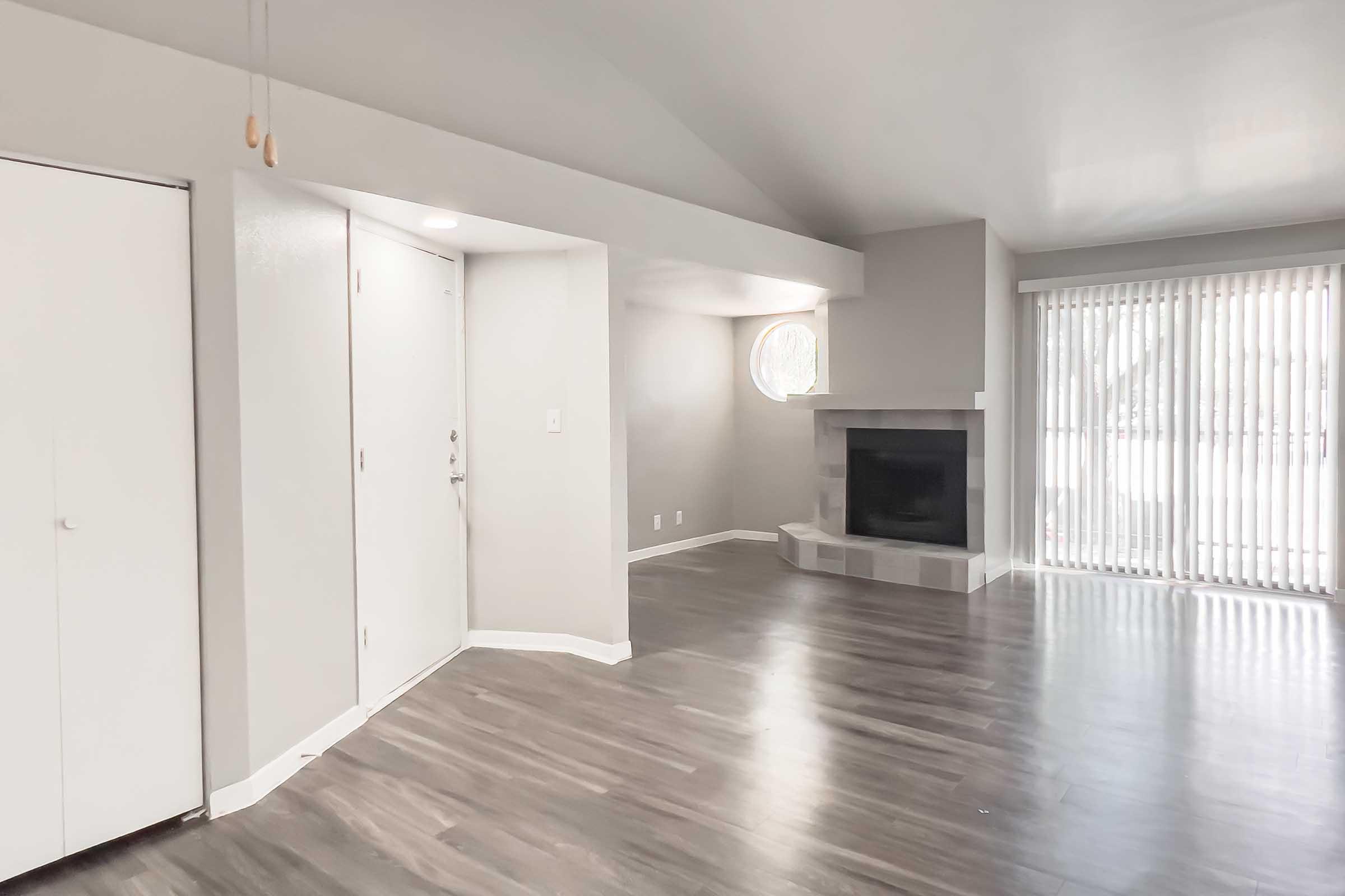Spacious interior of a modern living room featuring a fireplace, large windows with blinds, and a built-in closet. Neutral-colored walls and a high ceiling create an airy atmosphere. The flooring is dark wood, adding warmth to the space. Natural light streams in through the windows, enhancing the open feel.