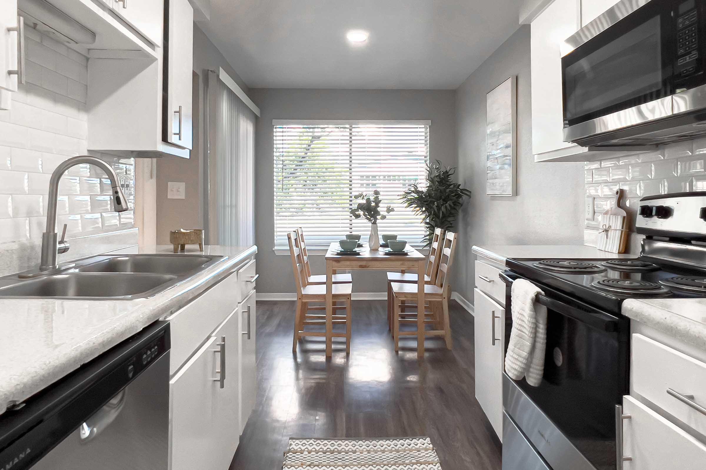 A modern kitchen featuring white cabinetry, a sink, and stainless steel appliances. Sunlight streams through a window with blinds, illuminating a small dining area with a table set for two. The floor is dark wood, and a decorative plant adds a touch of greenery.