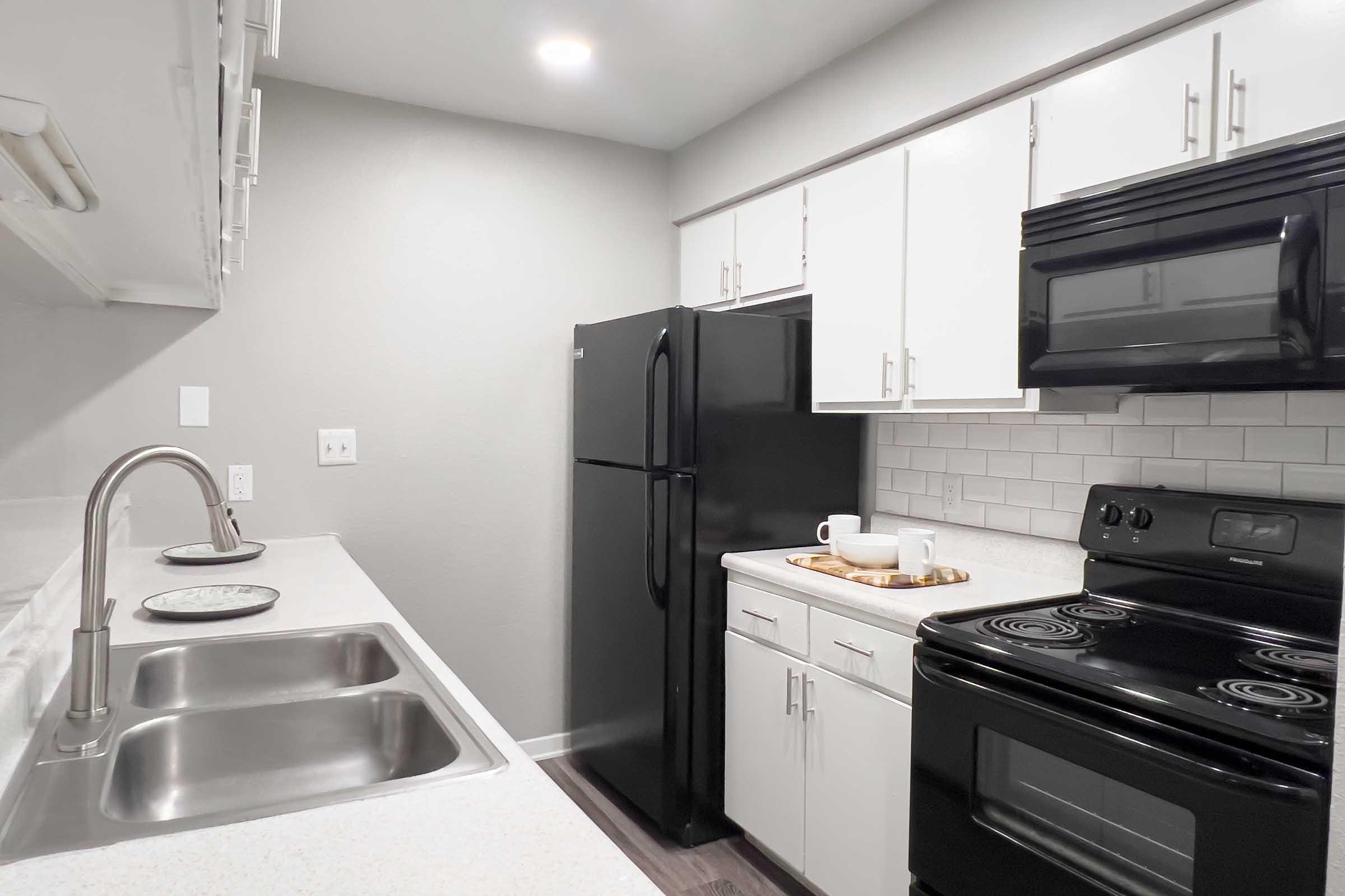 Modern kitchen featuring stainless steel sinks, a black refrigerator, and an electric stove. The cabinetry is white, complemented by a gray wall. A wooden tray with tableware is on the counter next to the stove, and there is under-cabinet lighting for added brightness.