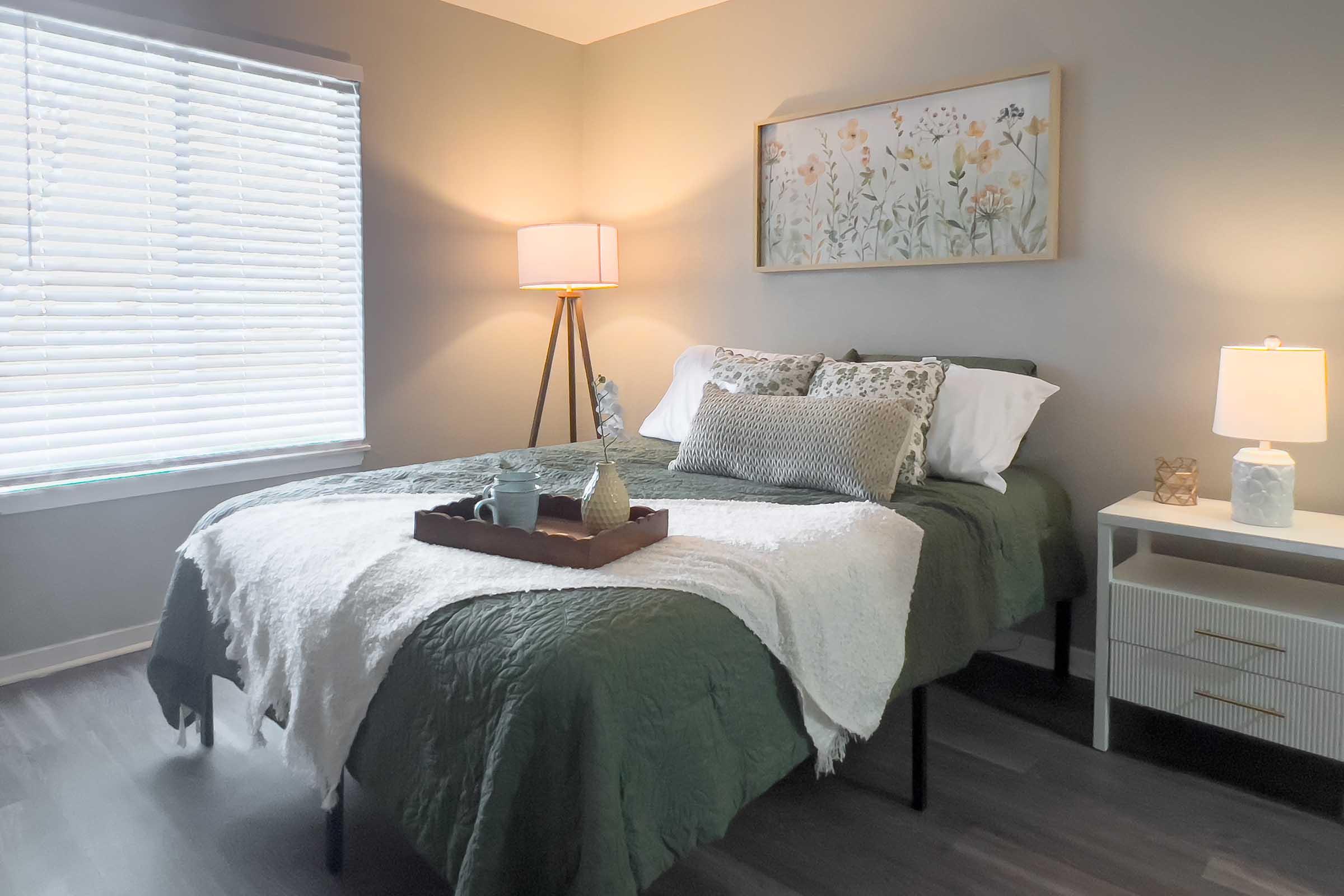 A cozy bedroom featuring a neatly made bed with a green comforter and decorative pillows. A tray on the bed holds small decorative items. Natural light filters through a window with white blinds, and there are two bedside lamps, one with a warm glow. A floral artwork is hung above the bed, enhancing the serene atmosphere.