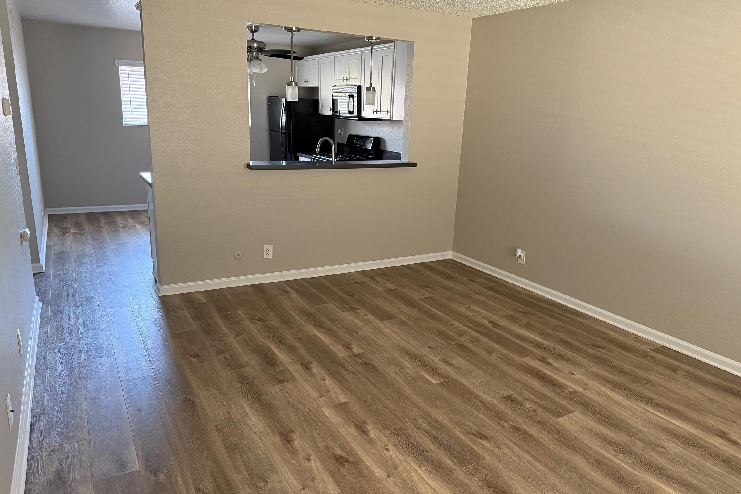 A spacious, empty room featuring laminate flooring, light-colored walls, and a large window. There is an open doorway leading to a kitchen area that is visible in the background. The kitchen includes modern appliances and cabinetry, enhancing the inviting atmosphere of the living space.