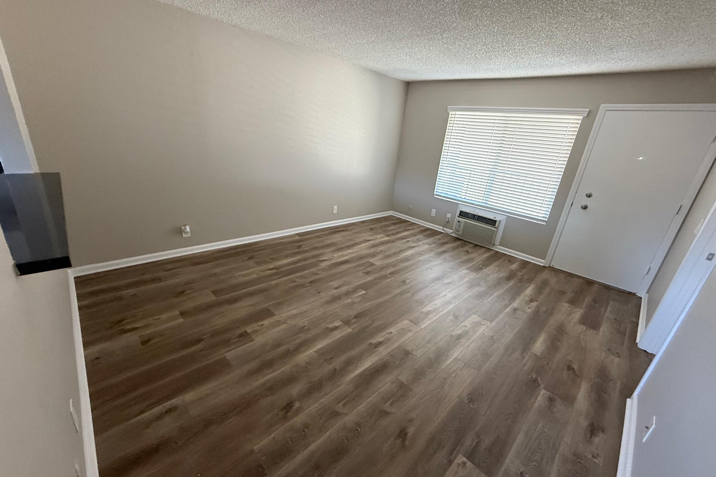 A spacious, empty living room with light-colored walls and a textured ceiling. The wooden laminate flooring adds warmth, and there is a large window with blinds letting in natural light. An air conditioning unit is visible on the wall, and a door leads outside.