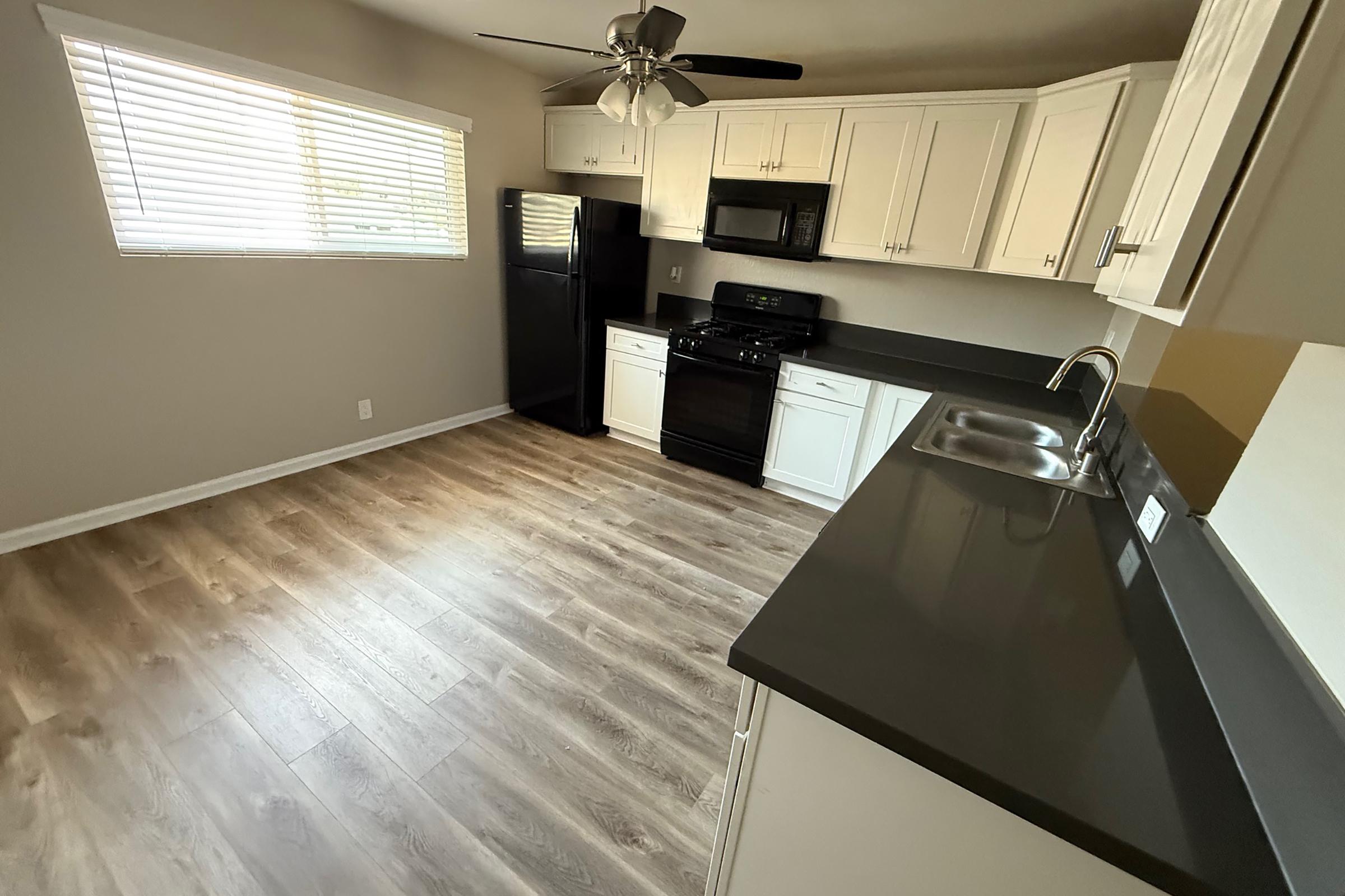 A modern kitchen featuring light-colored cabinetry and dark countertops. It includes a black refrigerator, a matching stove, and a sink with a stylish faucet. Natural light comes in through a window with blinds, illuminating the light wood-like flooring. The space is uncluttered and well-organized.