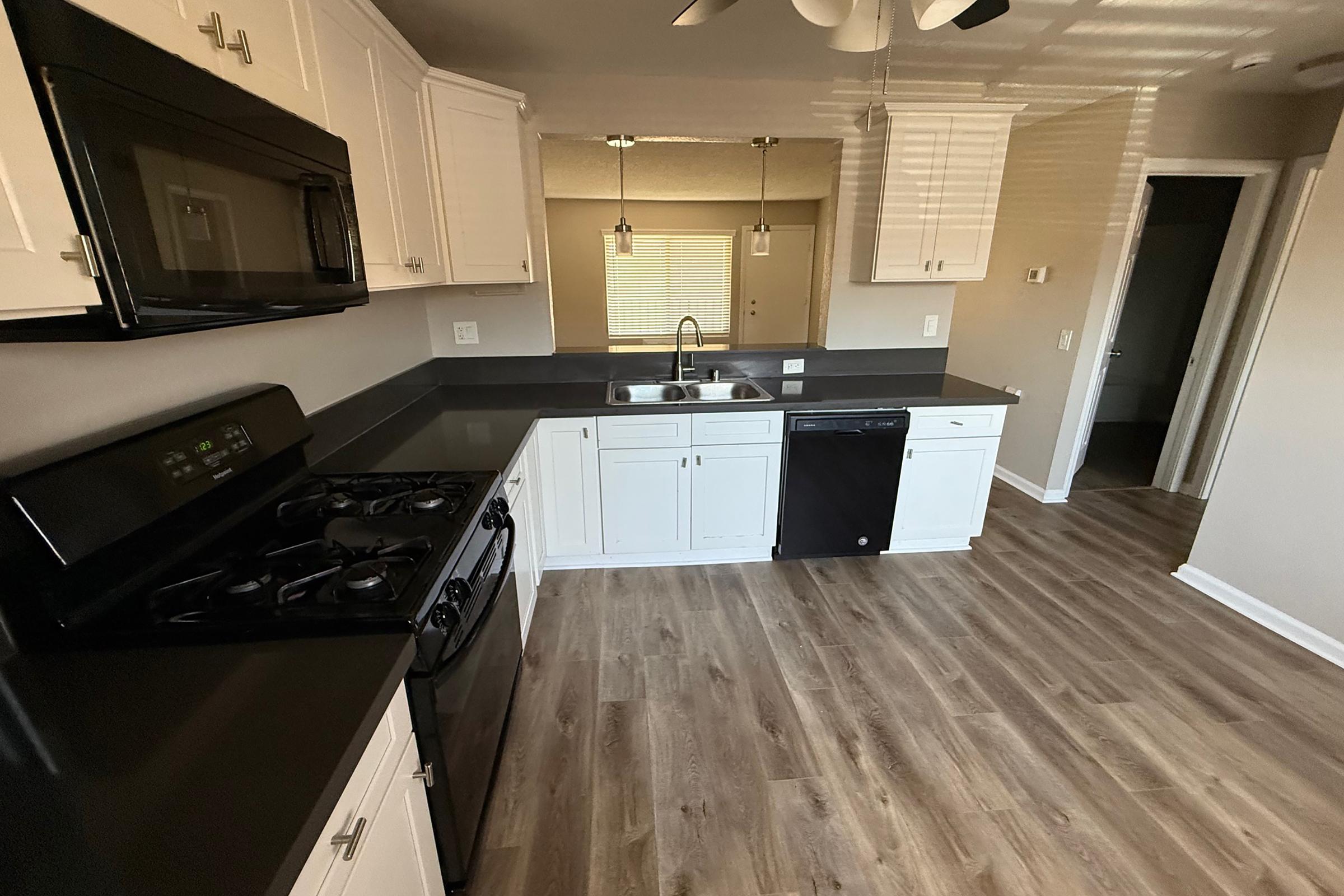 Modern kitchen featuring white cabinets, black countertops, and stainless steel appliances. The layout includes a gas stove, a sink, and a dishwasher. Natural light filters in through a window, with wooden floors adding warmth to the space. The kitchen connects to a hallway leading to another room.
