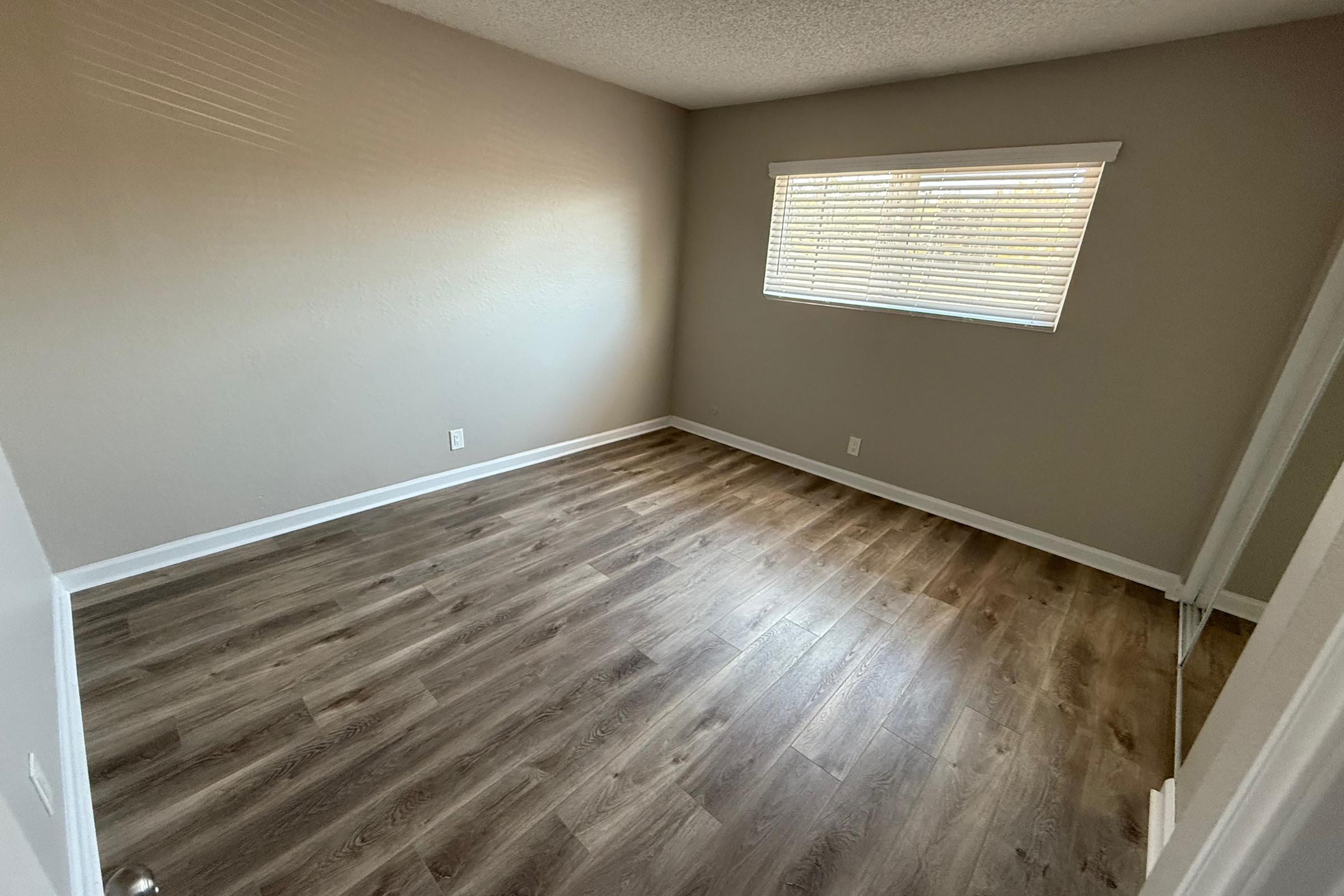 Empty room with a light-colored wall and wood-like laminate flooring. A window with blinds allows natural light in, illuminating the space. The room is devoid of furniture, creating a spacious and clean appearance, with a doorway visible on one side.