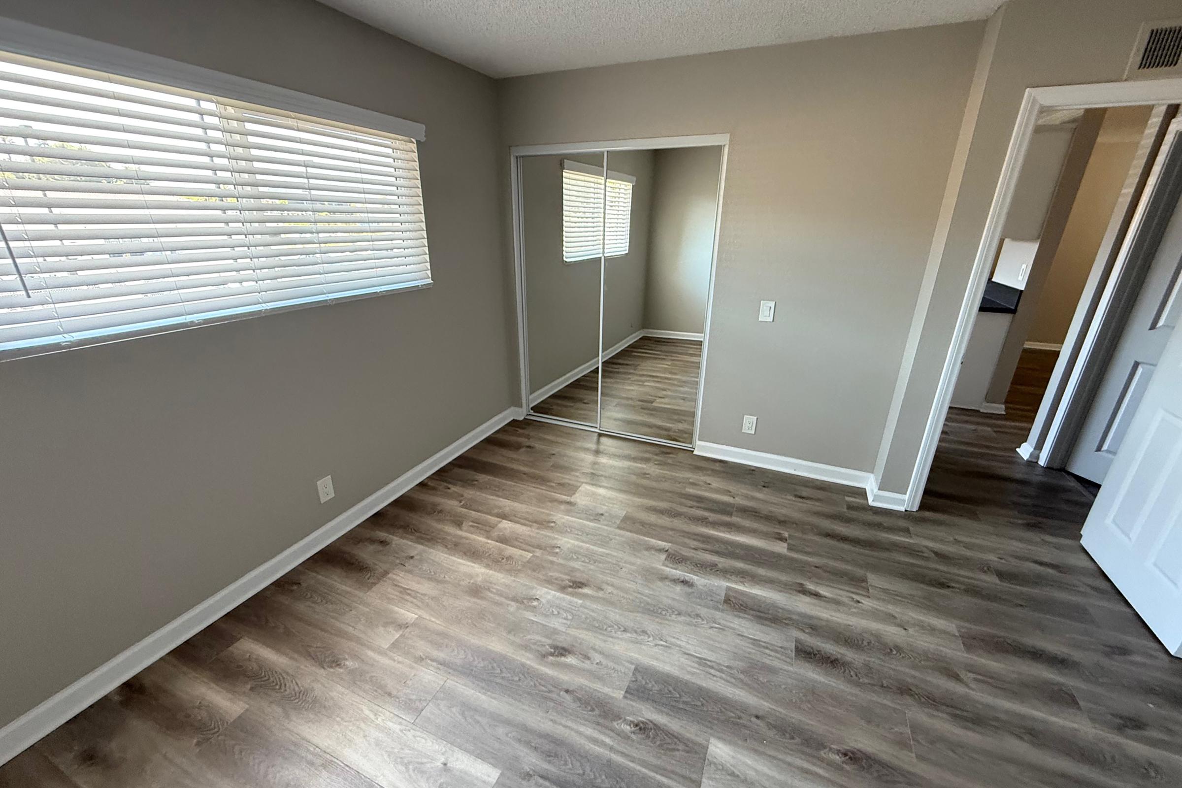 Interior view of a room featuring light-colored walls, a window with white blinds, and mirrored closet doors. The floor is made of wood-like laminate, and there is a door leading to another room. Natural light fills the space, creating a bright and inviting atmosphere.