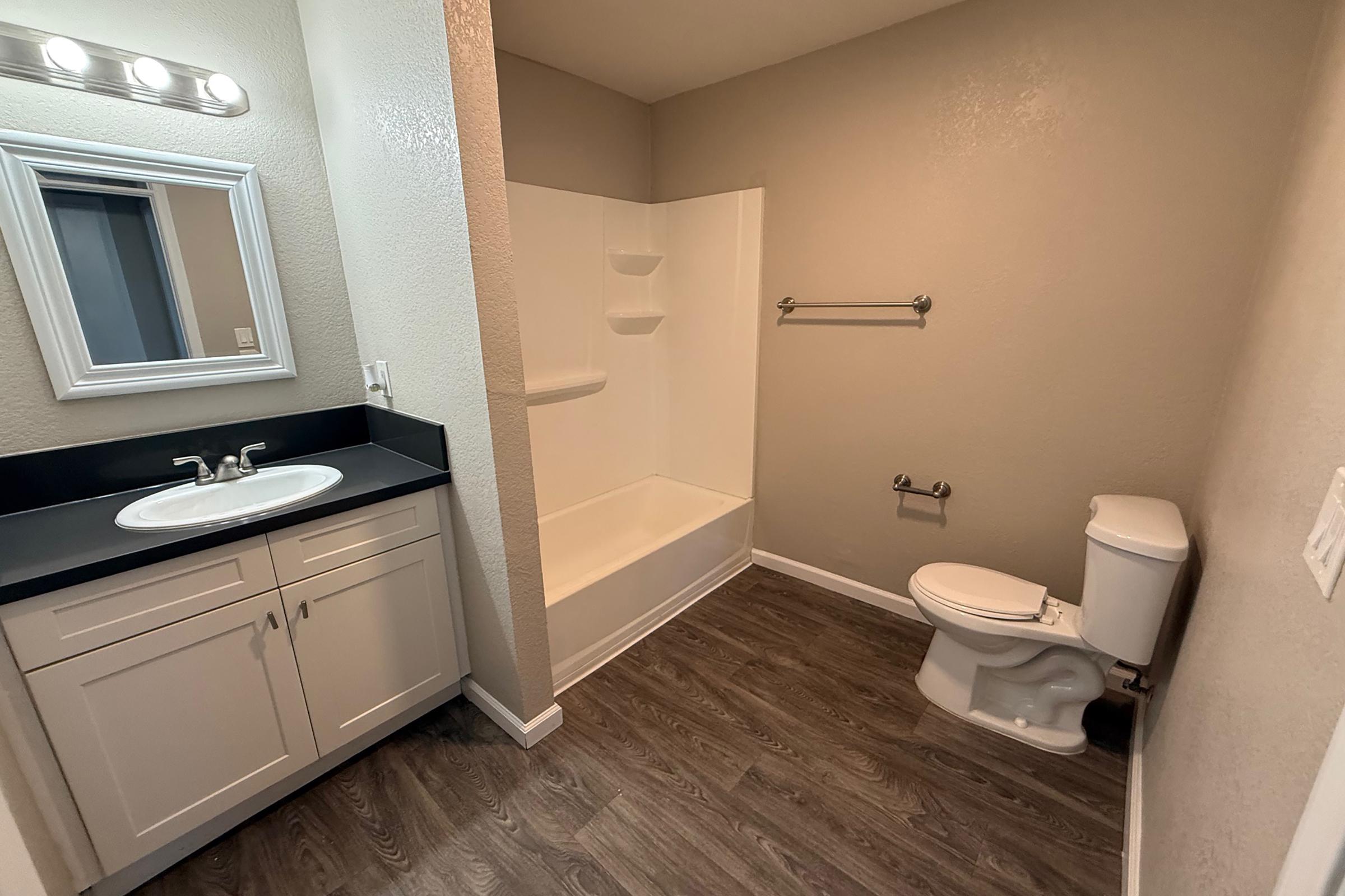 A bathroom featuring a white bathtub and shower, a toilet, a large mirror above a dark countertop with sink and cabinet below, and a towel bar mounted on the wall. The walls are painted in a light beige color, with a wood-like textured floor.