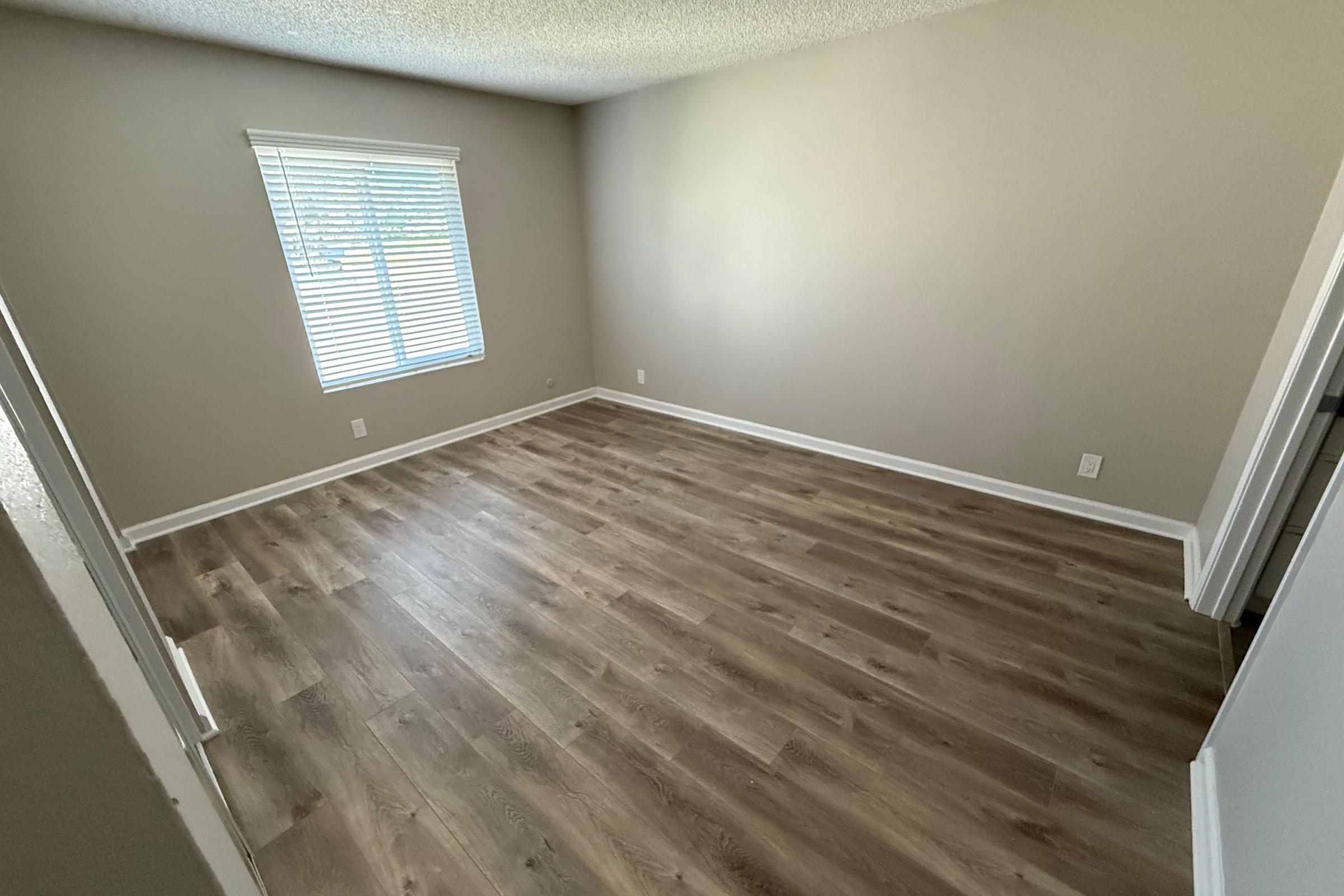 A spacious, empty room with light brown laminate flooring. A window with white blinds is on the left, allowing natural light to enter. The walls are painted in a neutral beige color, creating a bright, airy atmosphere. There are no furnishings in the room, emphasizing its open layout.