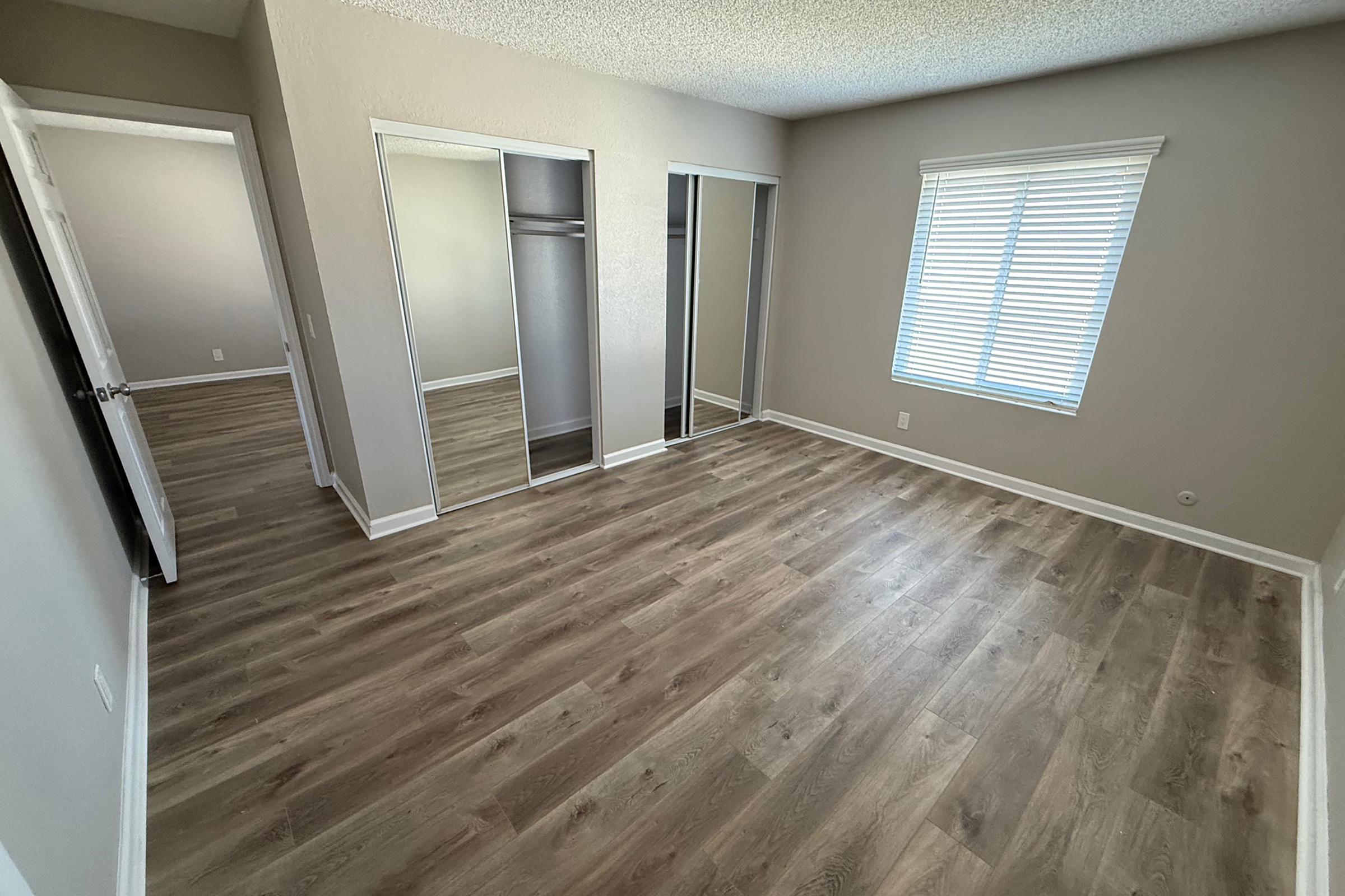 Empty room featuring a light wood floor, two mirrored closet doors on the left, and a window with blinds on the right. The walls are painted a light color, and there is a doorway visible leading to another space. The room is well-lit, suggesting a fresh and clean environment.